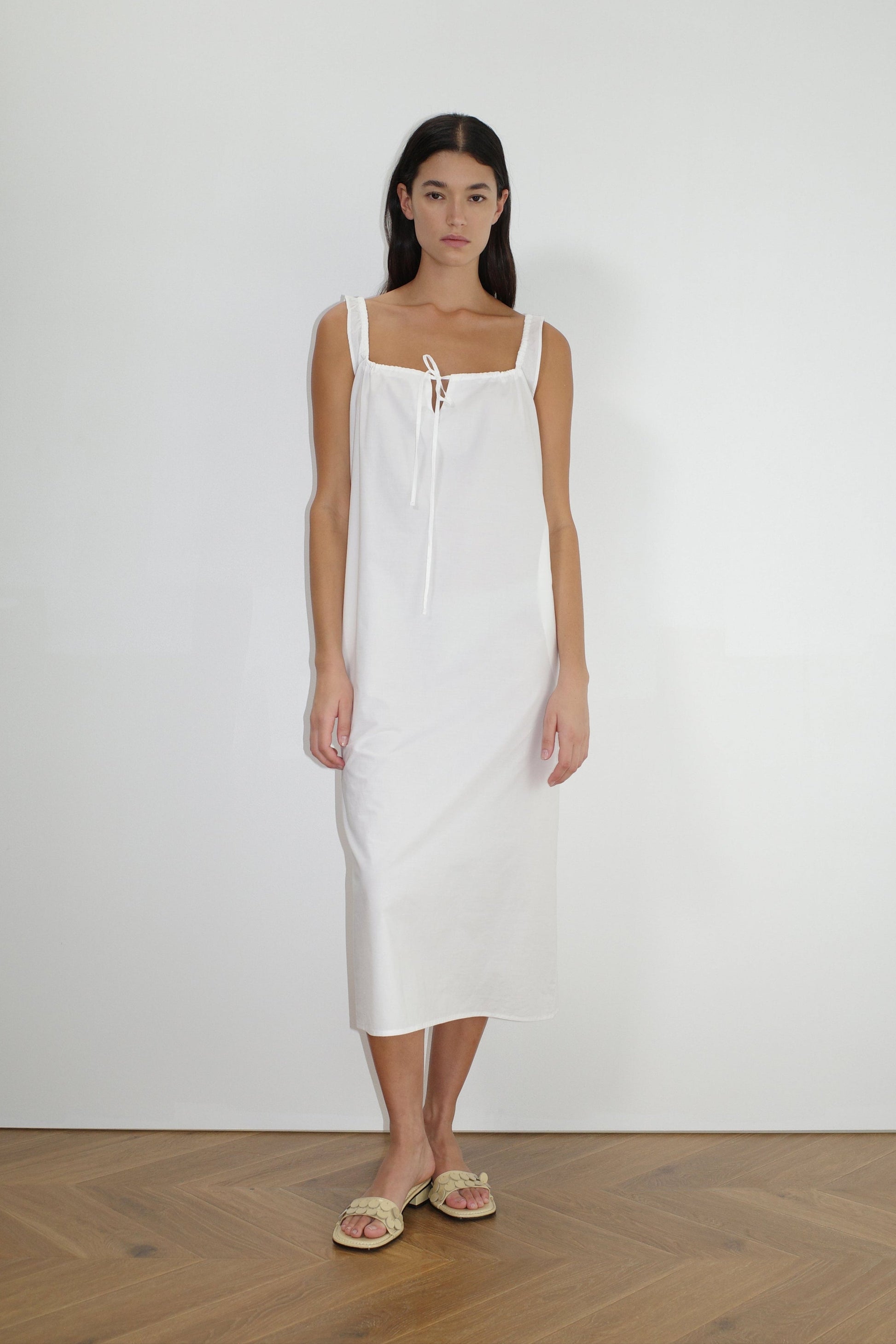 A woman with long dark hair and a neutral expression stands indoors against a plain white wall, wearing The Paper Dress - White by Deiji Studios, a sleeveless square-neck maxi dress with tie details, paired with light-colored sandals.