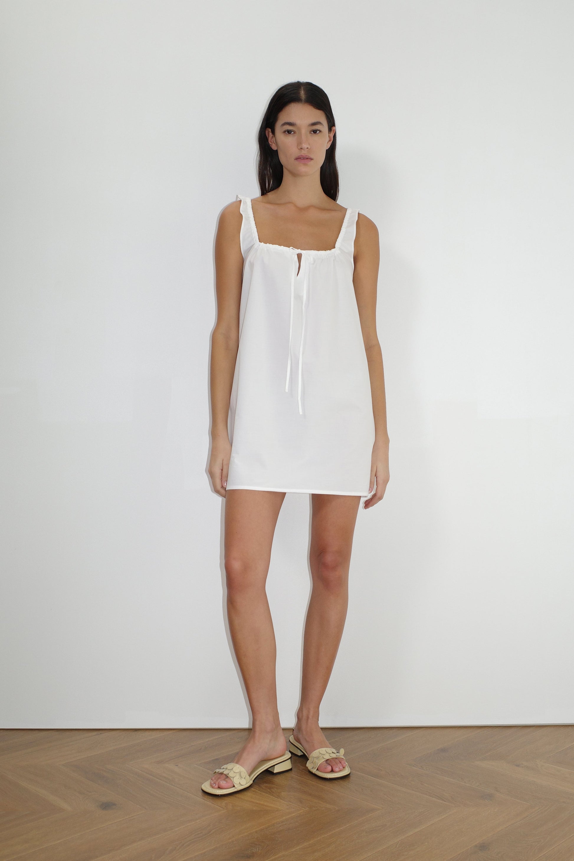 A woman stands indoors against a plain white wall, wearing The Bow Dress - White by Deiji Studios with a tie neckline and beige sandals. She has long dark hair, a neutral expression, and wooden flooring is visible.
