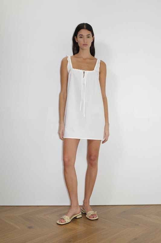 A woman stands indoors against a plain white wall, wearing The Bow Dress - White by Deiji Studios with a tie neckline and beige sandals. She has long dark hair, a neutral expression, and wooden flooring is visible.