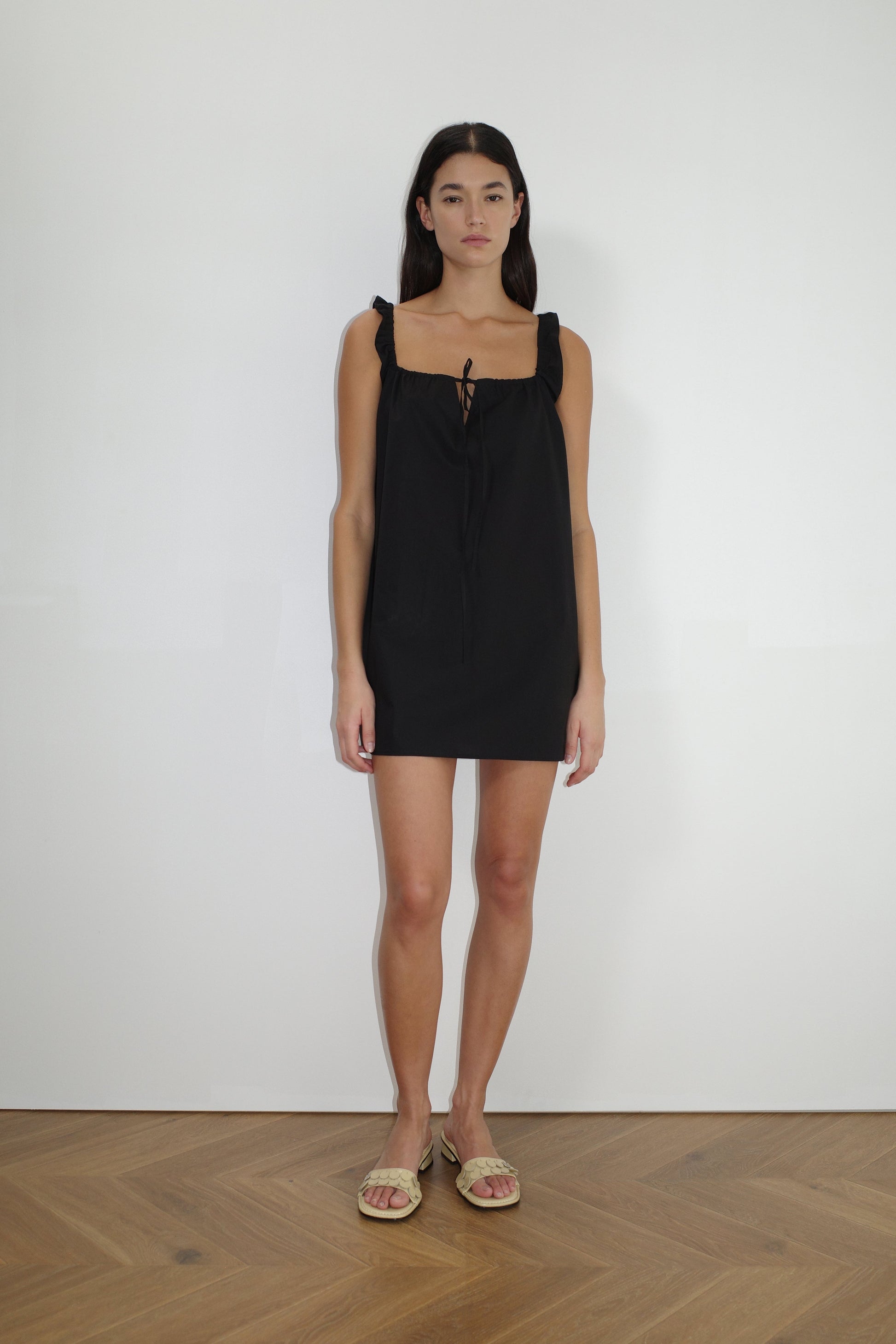 A woman with long dark hair stands against a plain white wall, wearing The Bow Dress - Black by Deiji Studios with ruffled straps and a bow at the neckline, paired with beige flat sandals on a light wood floor.