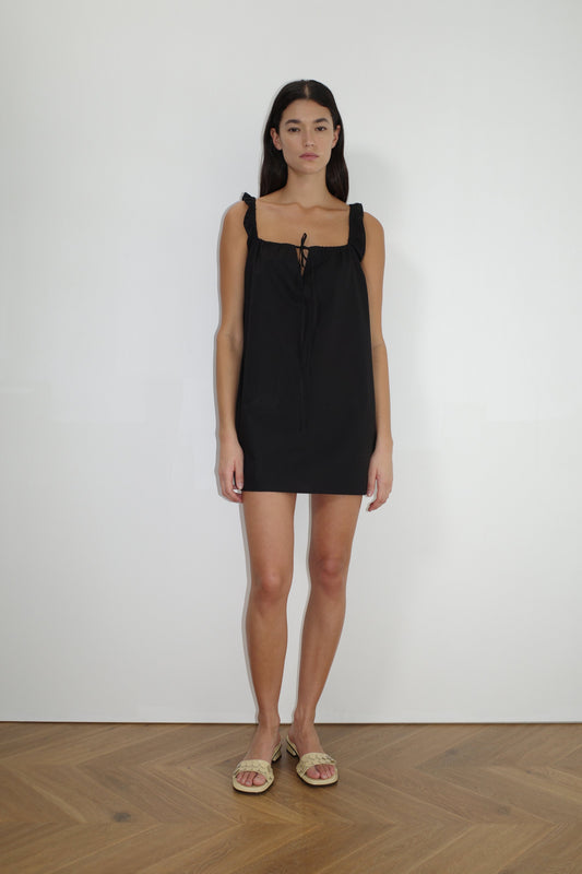 A woman with long dark hair stands against a plain white wall, wearing The Bow Dress - Black by Deiji Studios with ruffled straps and a bow at the neckline, paired with beige flat sandals on a light wood floor.