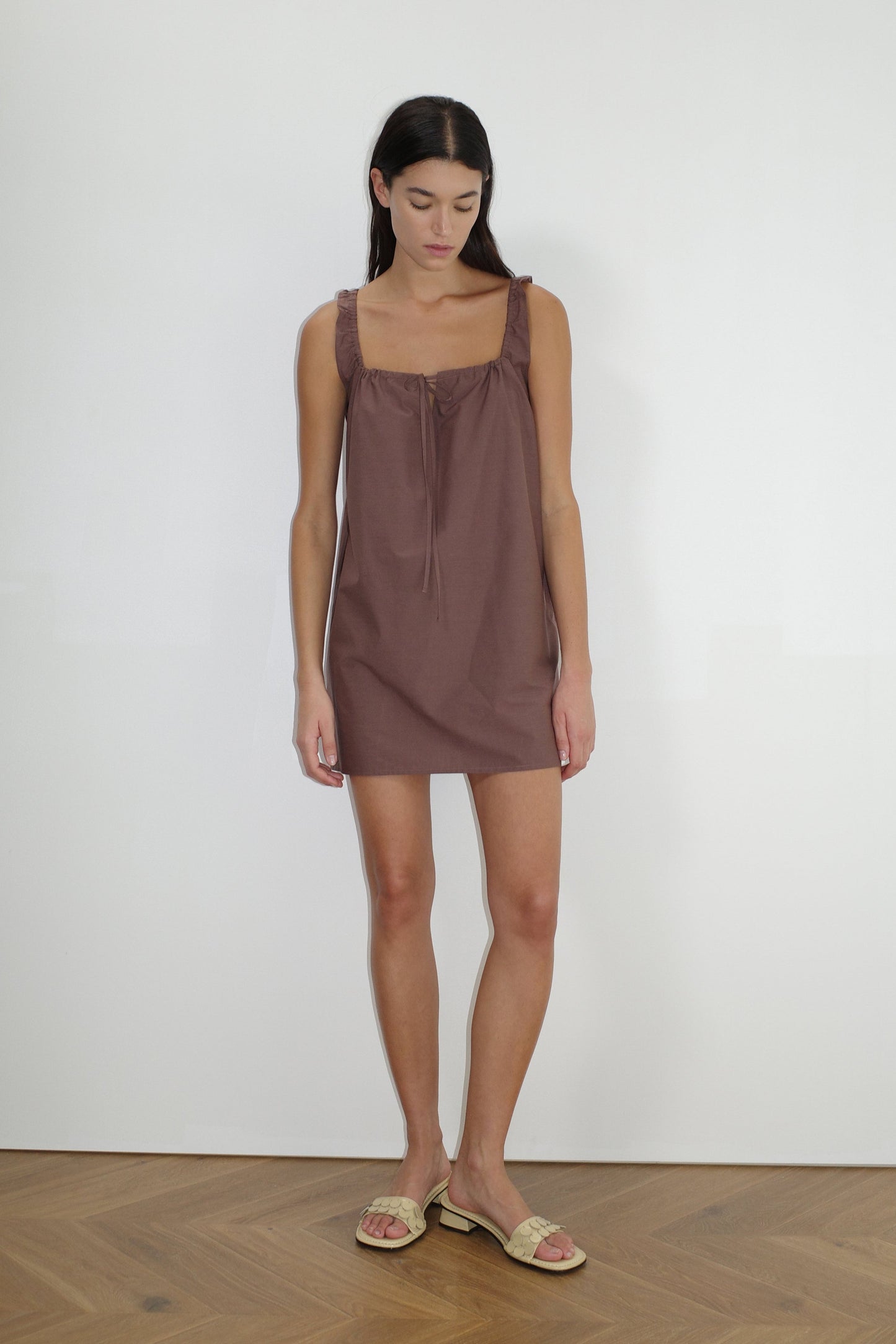 A woman stands indoors against a plain white wall, wearing The Bow Dress - Cedar, a mini-length dress with a gathered neckline and tie detail, paired with beige slide sandals. She has long dark hair and a neutral expression.