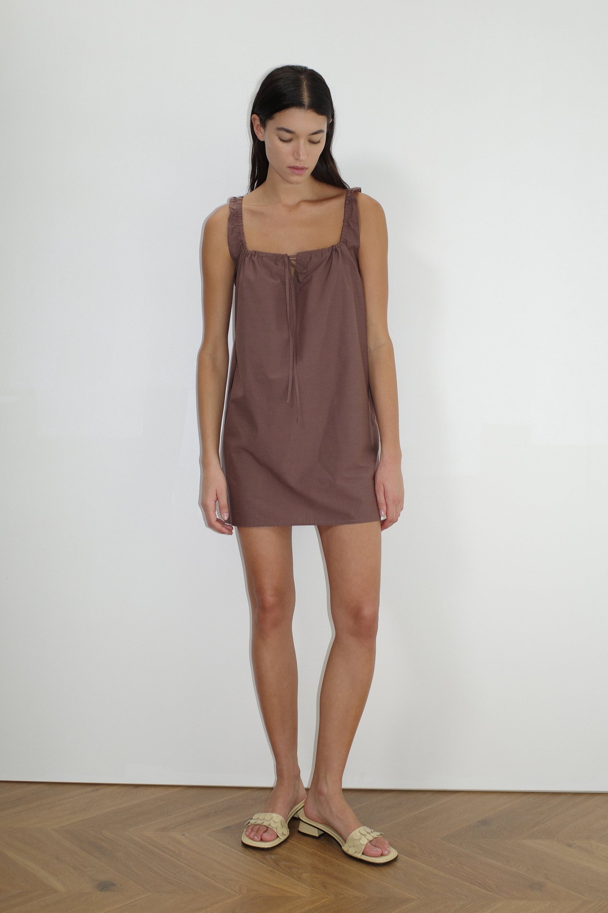 A woman stands indoors against a plain white wall, wearing The Bow Dress - Cedar, a mini-length dress with a gathered neckline and tie detail, paired with beige slide sandals. She has long dark hair and a neutral expression.