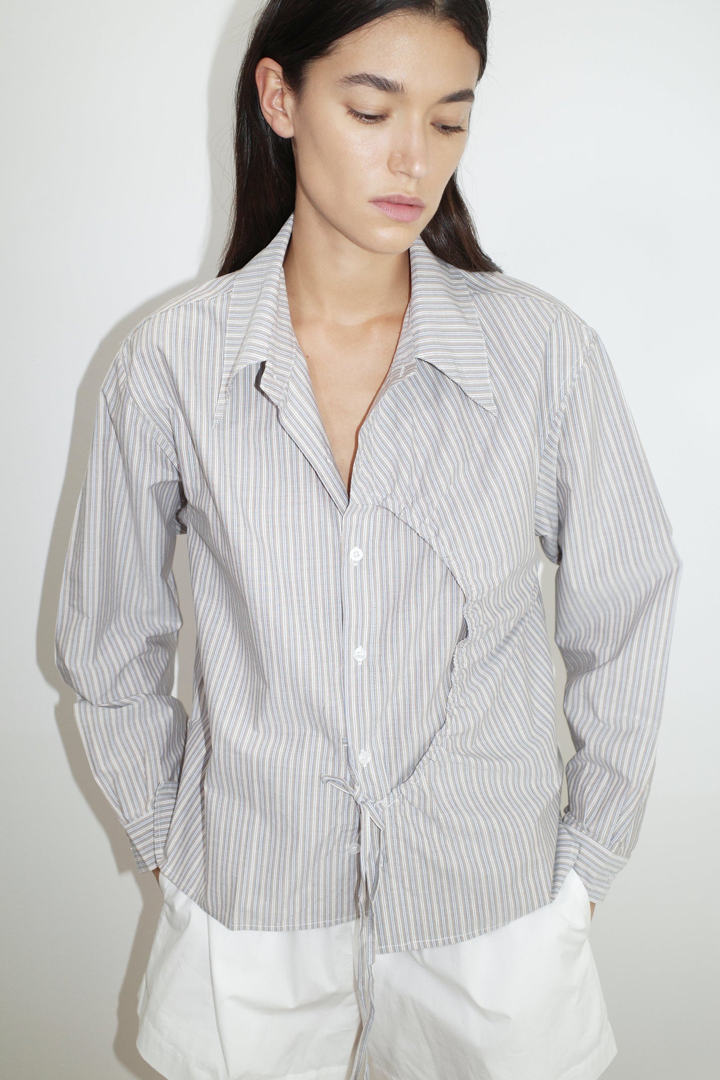 A woman with long dark hair wears the Deiji Studios Arch Tie Shirt - Seas Stripe, a light blue and white organic cotton top with an irregular circular seam, standing against a plain white background with her hands in her white skirt pockets.