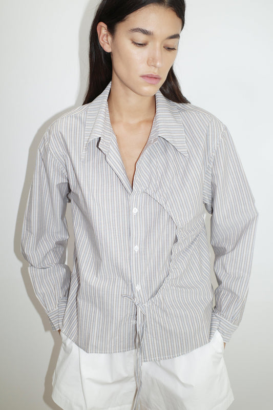 A woman with long dark hair wears the Deiji Studios Arch Tie Shirt - Seas Stripe, a light blue and white organic cotton top with an irregular circular seam, standing against a plain white background with her hands in her white skirt pockets.