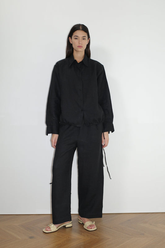 A person stands indoors against a white wall, wearing the Tie Split Set - Black with an oversized button-up shirt and matching wide-leg pants. They have long dark hair, beige flat sandals, and stand on a wooden floor.