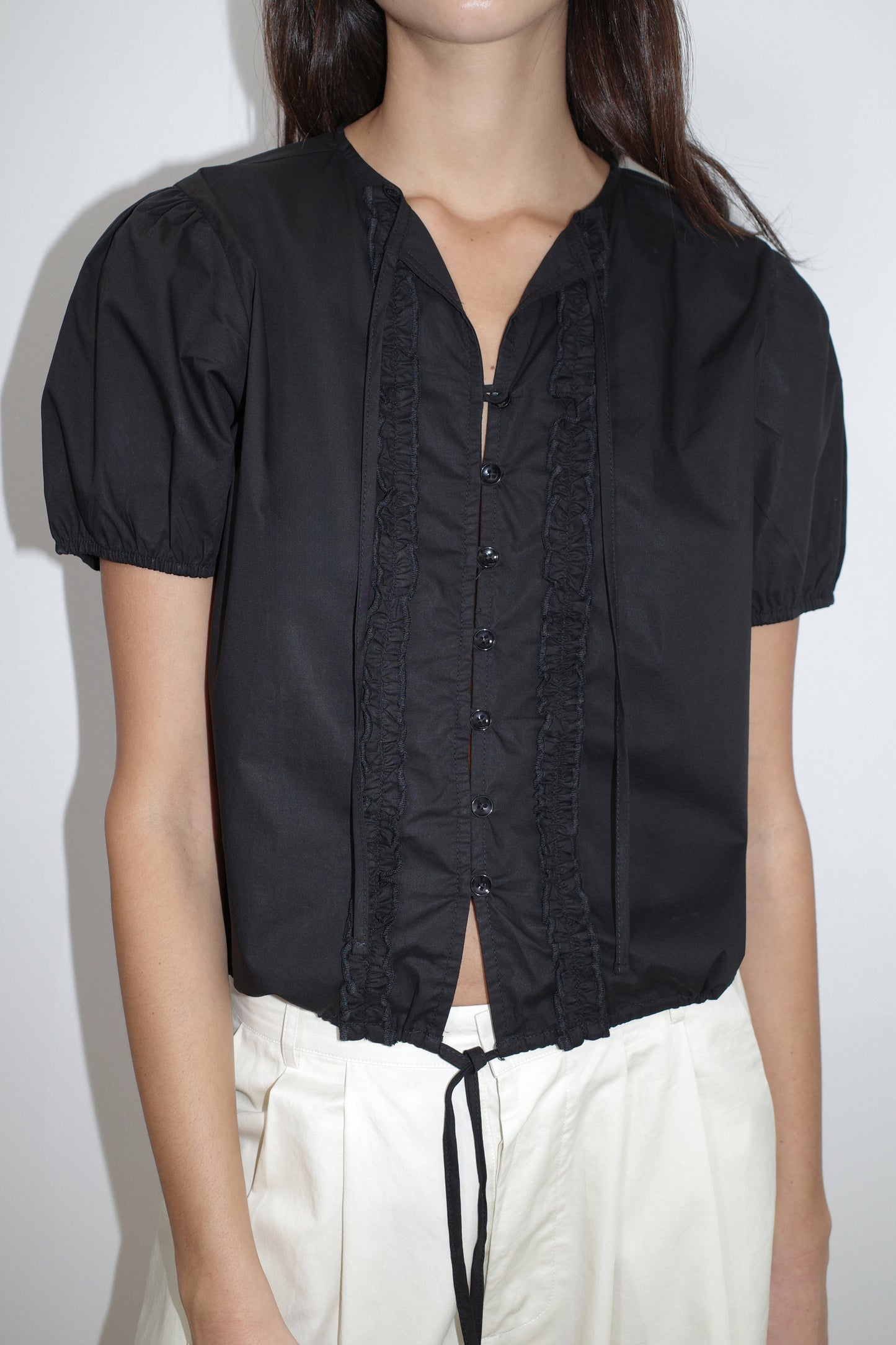 A person wears the Deiji Studios Double Frill Top - Black, a short-sleeved blouse with front buttons, paired with high-waisted loose white pants. The background is plain white.