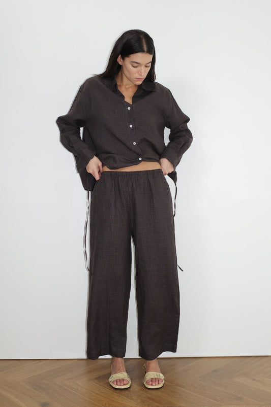 A woman stands indoors against a white wall, wearing the Tie Split Set—a loose, dark brown button-up shirt with matching wide-leg pants. She looks down with her hands in her pockets and has on flat beige sandals.