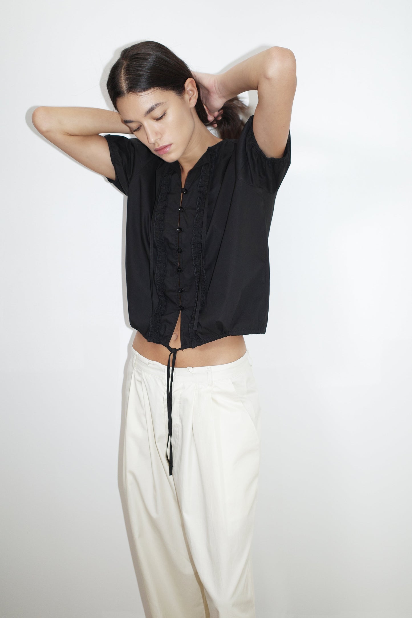 A person stands against a plain white background, wearing the Double Frill Top - Black by Deiji Studios and loose white pants, head tilted down with hands raised to adjust their hair.