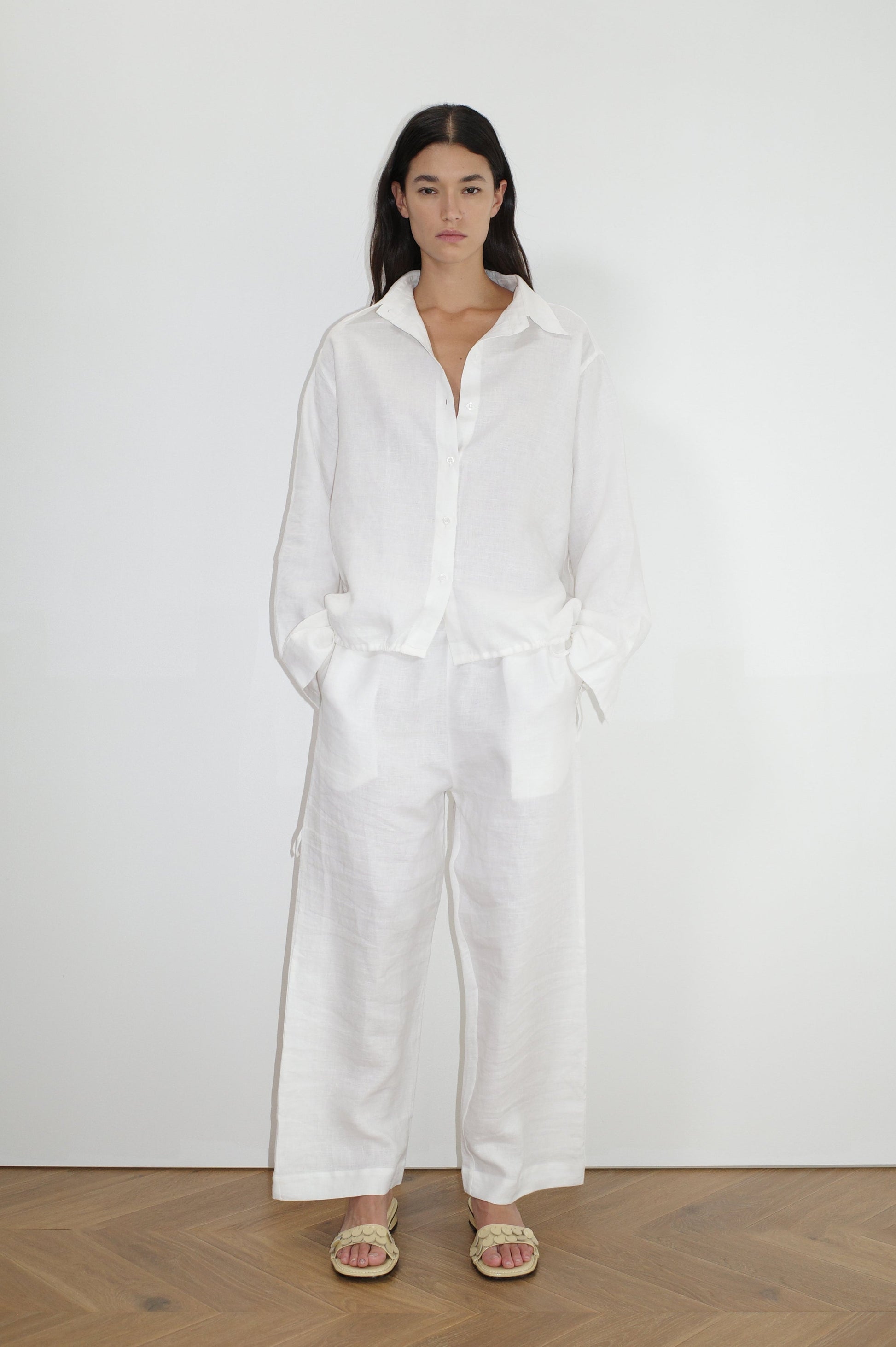 A woman with long dark hair stands indoors against a plain white wall, wearing the Tie Split Set—a loose-fitting white button-down shirt with matching wide-leg pants. She has her hands in her pockets and is also wearing sandals.