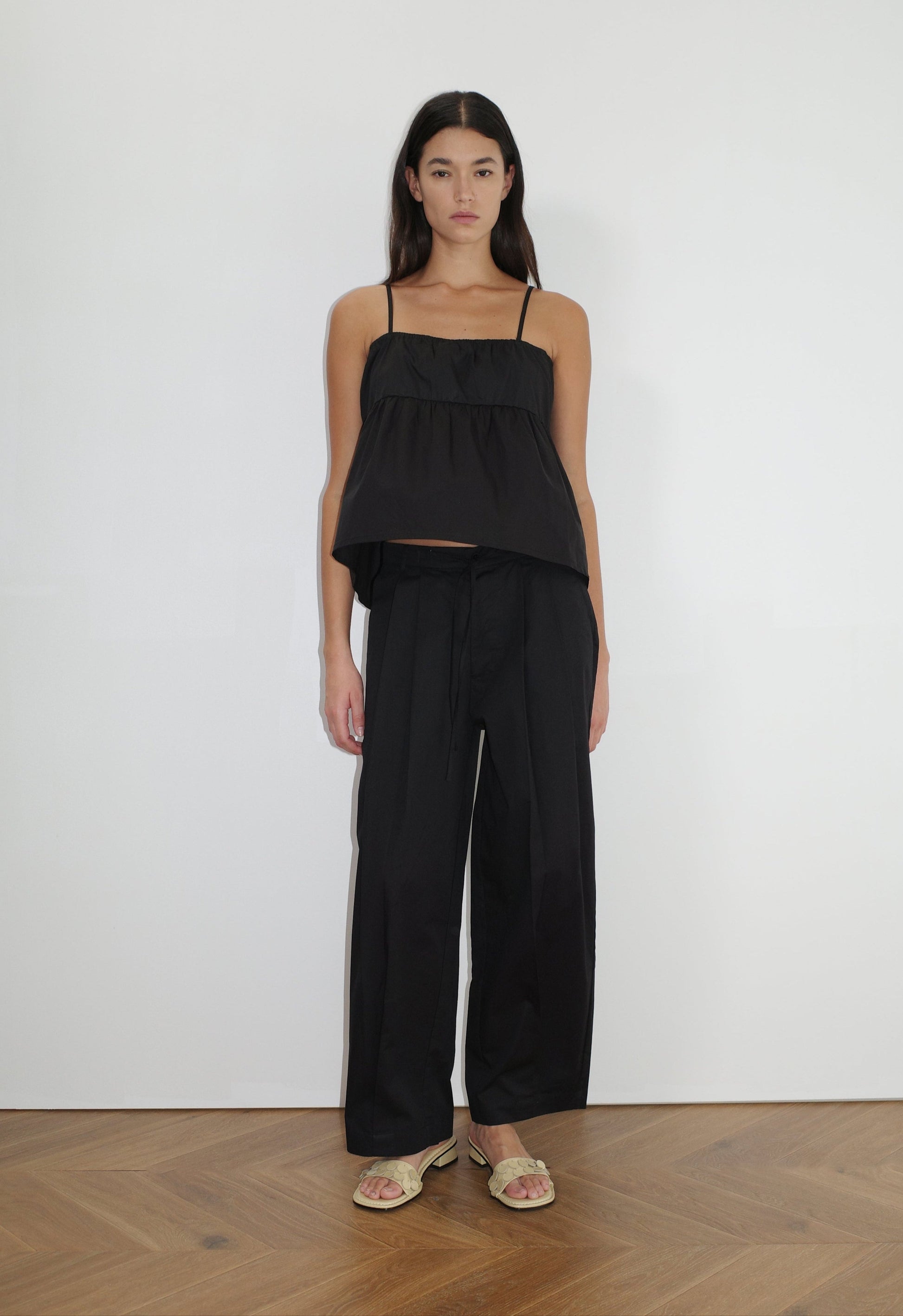 female model wears the turn pant in black, a relaxed tailored pant features soft pleating at the front, buttoned waistband and adjustable inner drawcord. designed with hidden side seam pockets and pockets at the back, made from 100% GOTS certified organic cotton paired with a black top