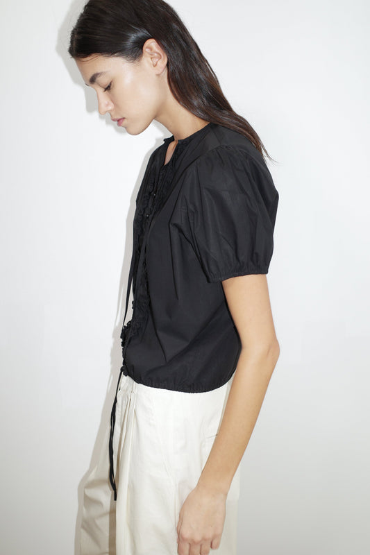 A woman with long dark hair, wearing the Deiji Studios Double Frill Top in black and high-waisted white pants, stands in profile against a plain white background, looking down with a neutral expression.
