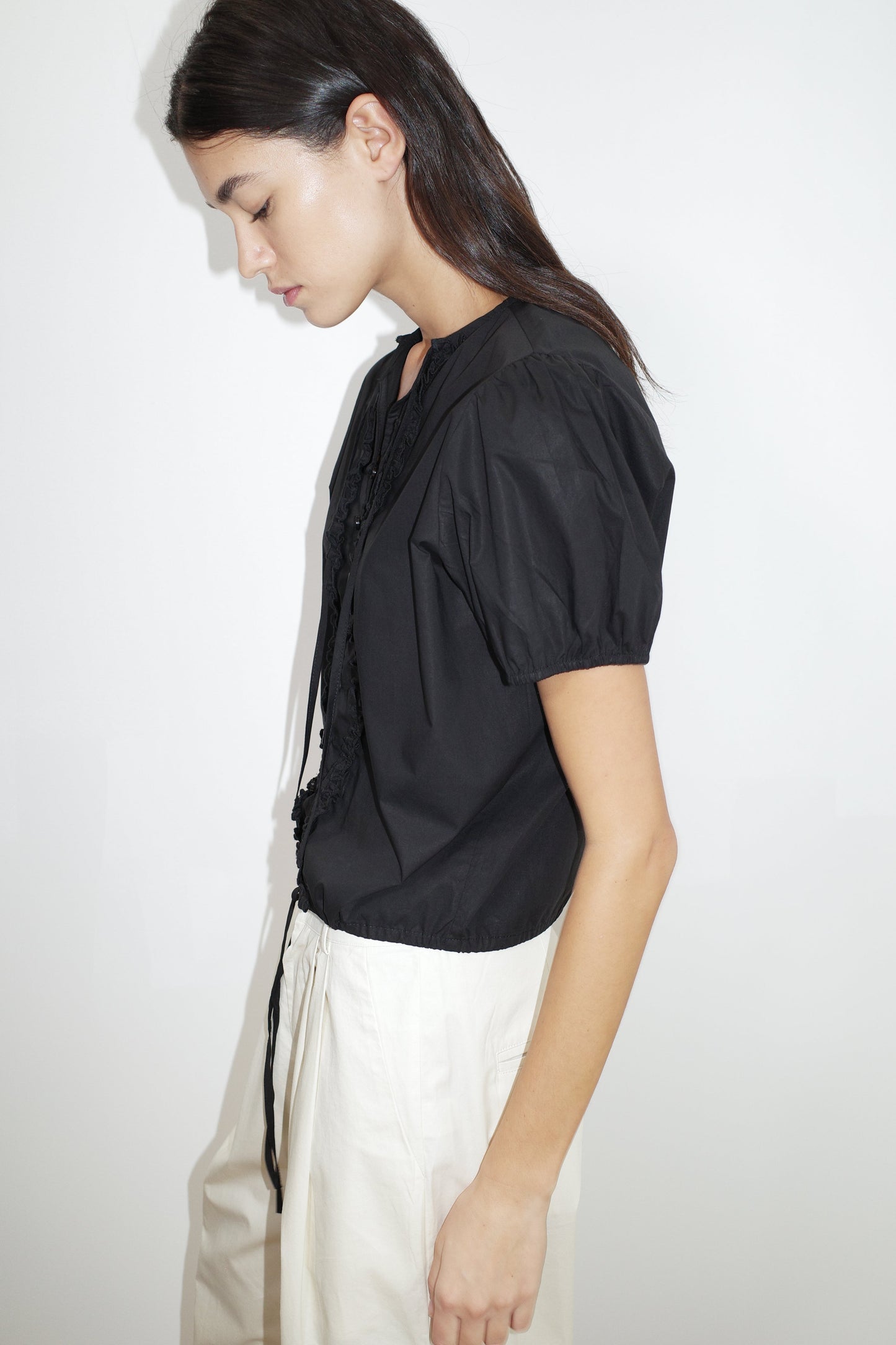 A woman with long dark hair, wearing the Deiji Studios Double Frill Top in black and high-waisted white pants, stands in profile against a plain white background, looking down with a neutral expression.