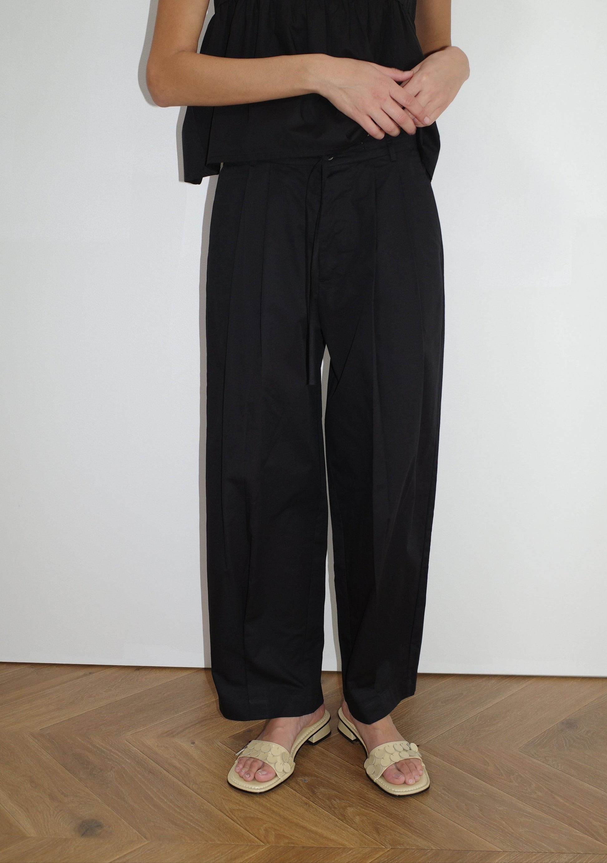 A person stands indoors on a wooden floor, wearing Deiji Studios' Turn Pant - Black with an adjustable drawcord, a black top, and light woven sandals. The upper part of their face is not visible.