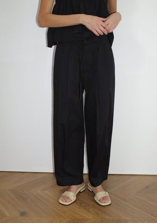 A person stands indoors on a wooden floor, wearing Deiji Studios' Turn Pant - Black with an adjustable drawcord, a black top, and light woven sandals. The upper part of their face is not visible.