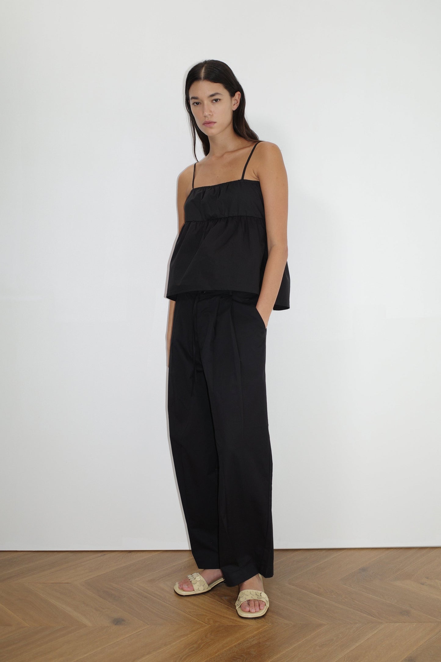 female model wears the turn pant in black, a relaxed tailored pant features soft pleating at the front, buttoned waistband and adjustable inner drawcord. designed with hidden side seam pockets and pockets at the back, made from 100% GOTS certified organic cotton paired with a black top