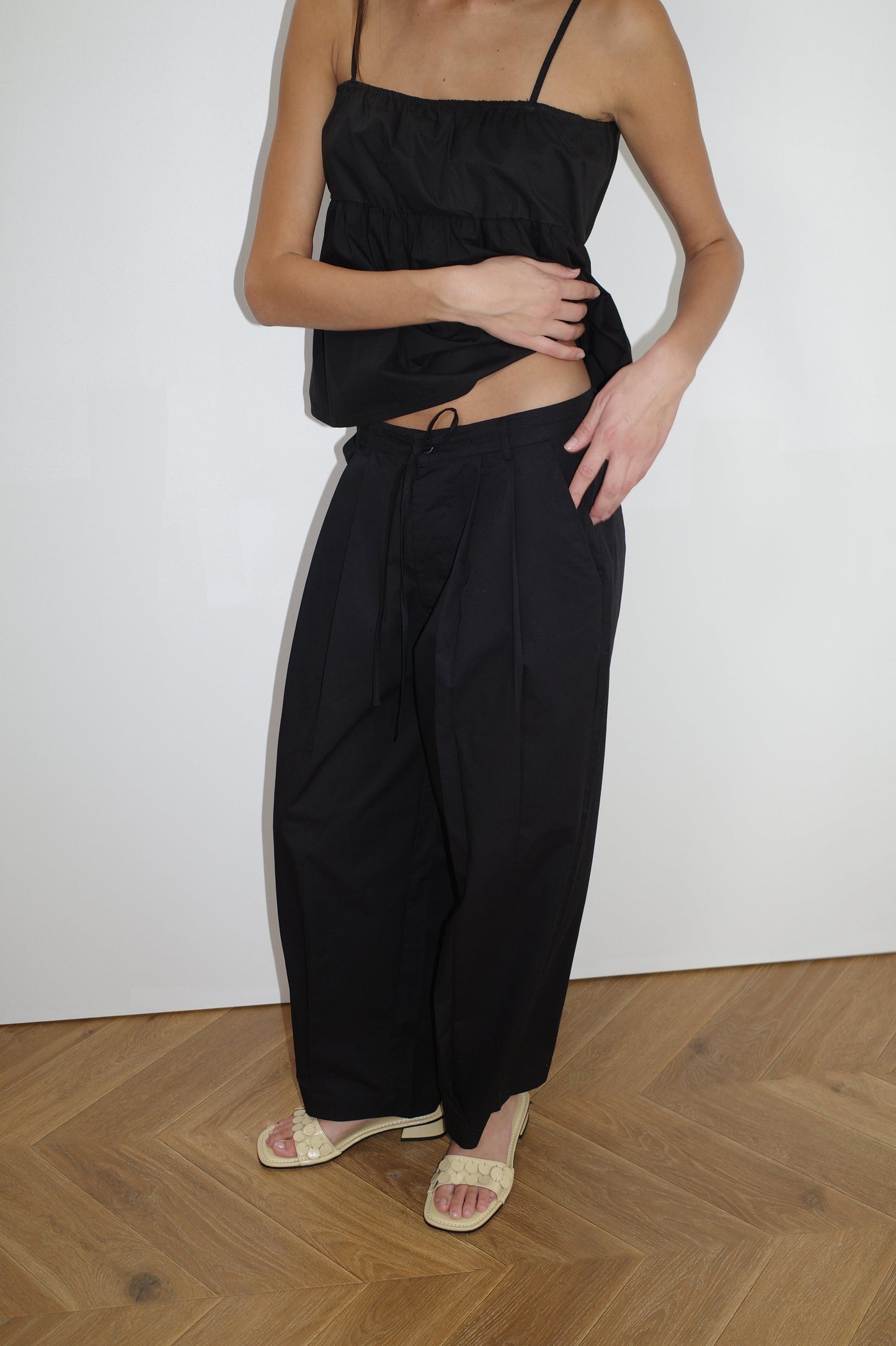 female model wears the turn pant in black, a relaxed tailored pant features soft pleating at the front, buttoned waistband and adjustable inner drawcord. designed with hidden side seam pockets and pockets at the back, made from 100% GOTS certified organic cotton paired with a black top