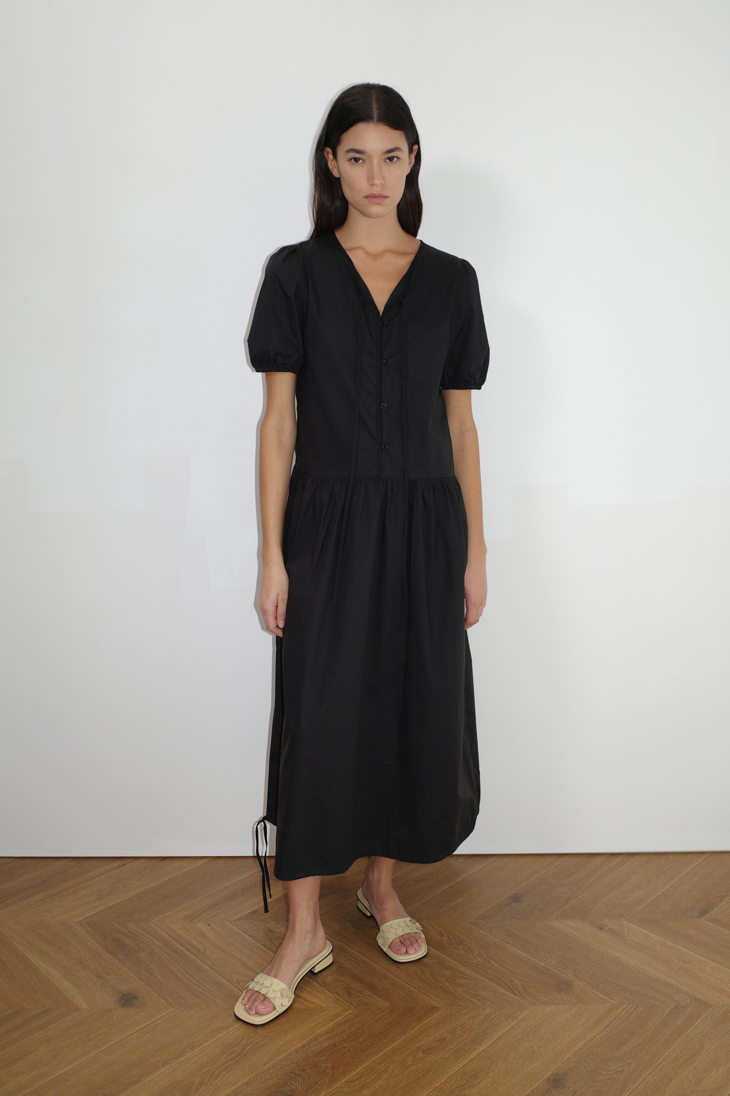 female model wears the draw hem dress by Deiji Studios in black


elevated everyday dress in mid length features a flattering v neckline with tie, button front detail with a drop waist, and soft elastic blouson sleeve


made from 100% organic cotton poplin