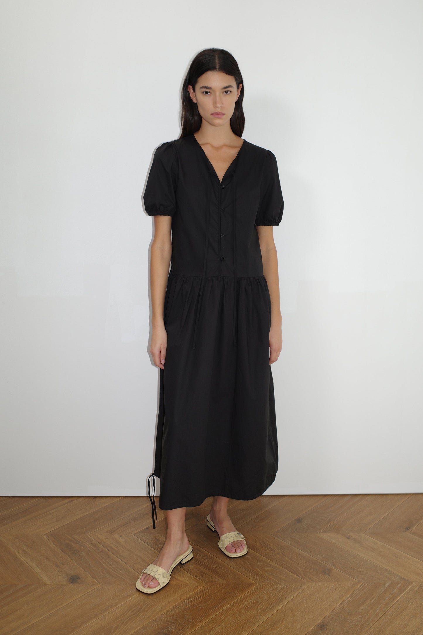 female model wears the draw hem dress by Deiji Studios in black


elevated everyday dress in mid length features a flattering v neckline with tie, button front detail with a drop waist, and soft elastic blouson sleeve


made from 100% organic cotton poplin