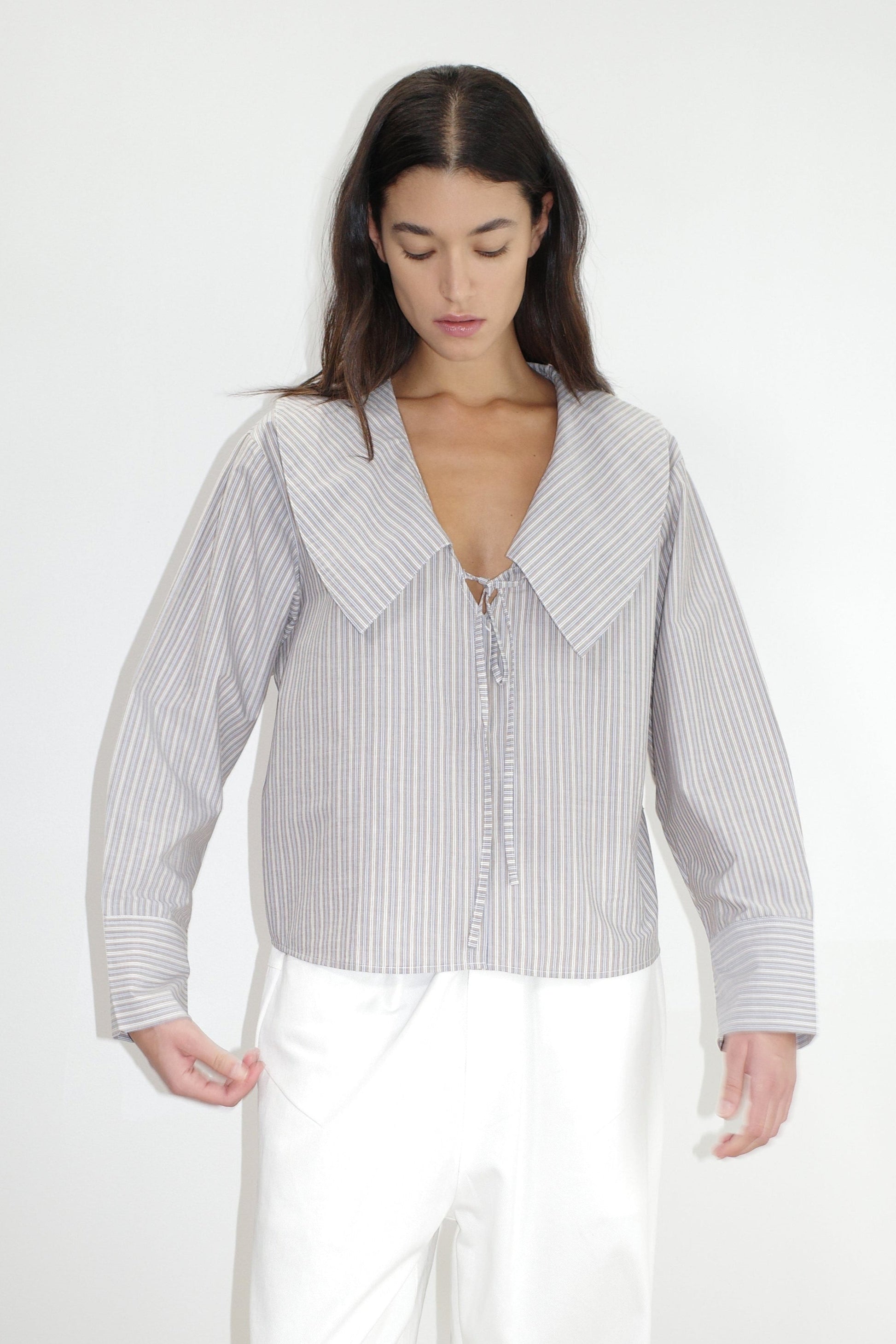female model wears the collared tie shirt by Deiji Studios in seas stripe


a structured shirt features an oversized collar and cuff, tie at front collar opening, dropped shoulder and elongated sleeves in a boxy fit


made from 100% GOTS certified organic cotton poplin

paired with a white cotton trouser