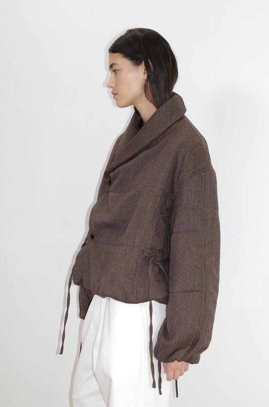 A person with straight, dark hair wears the oversized Bubble Coat - Herringbone by 0, featuring a high collar and drawstrings, paired with loose white pants against a plain white background.