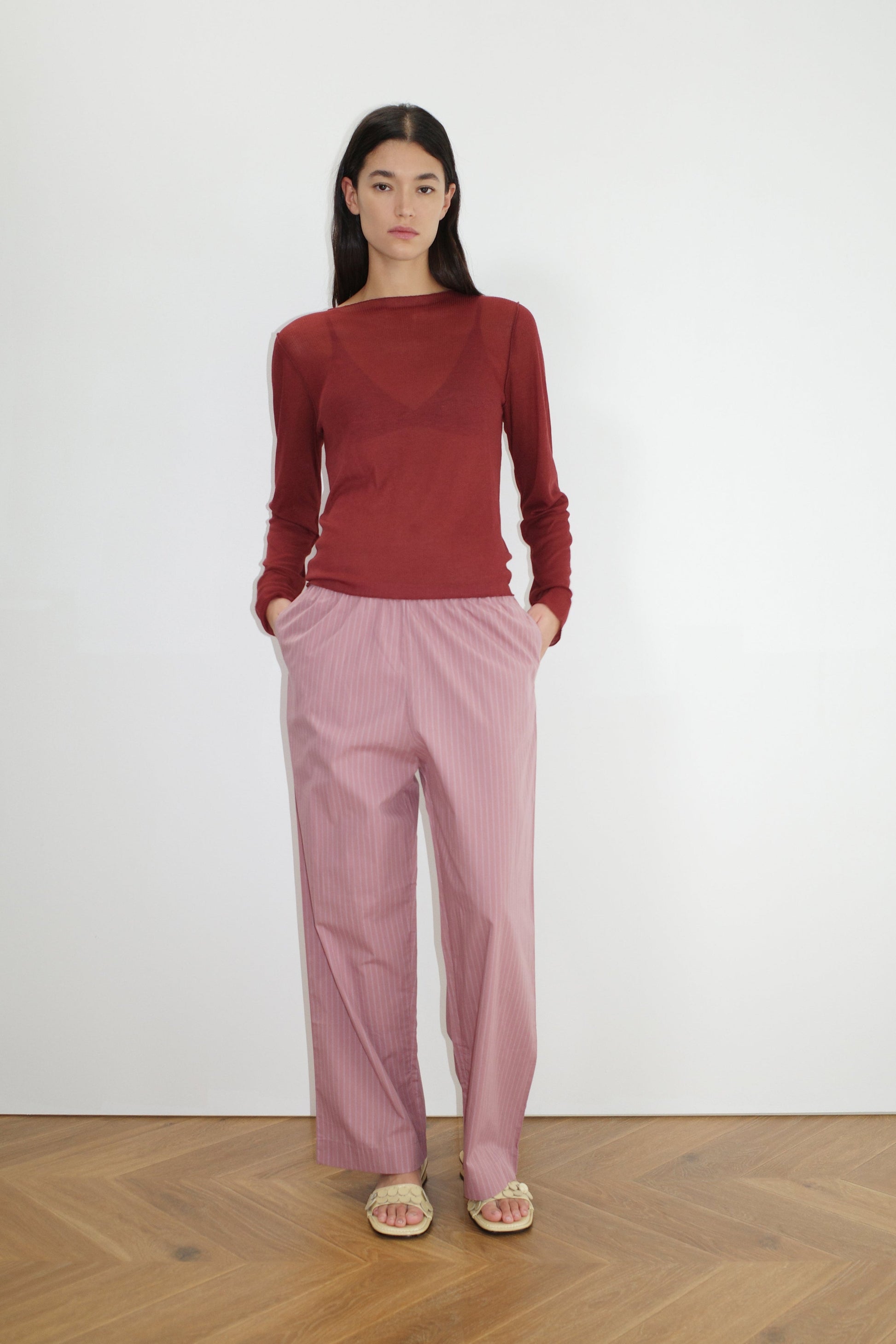 A woman stands against a white wall, hands in pockets, wearing loose pink pinstriped Ease Trouser, a sheer reddish-brown long-sleeved top, and beige sandals on a wooden floor.