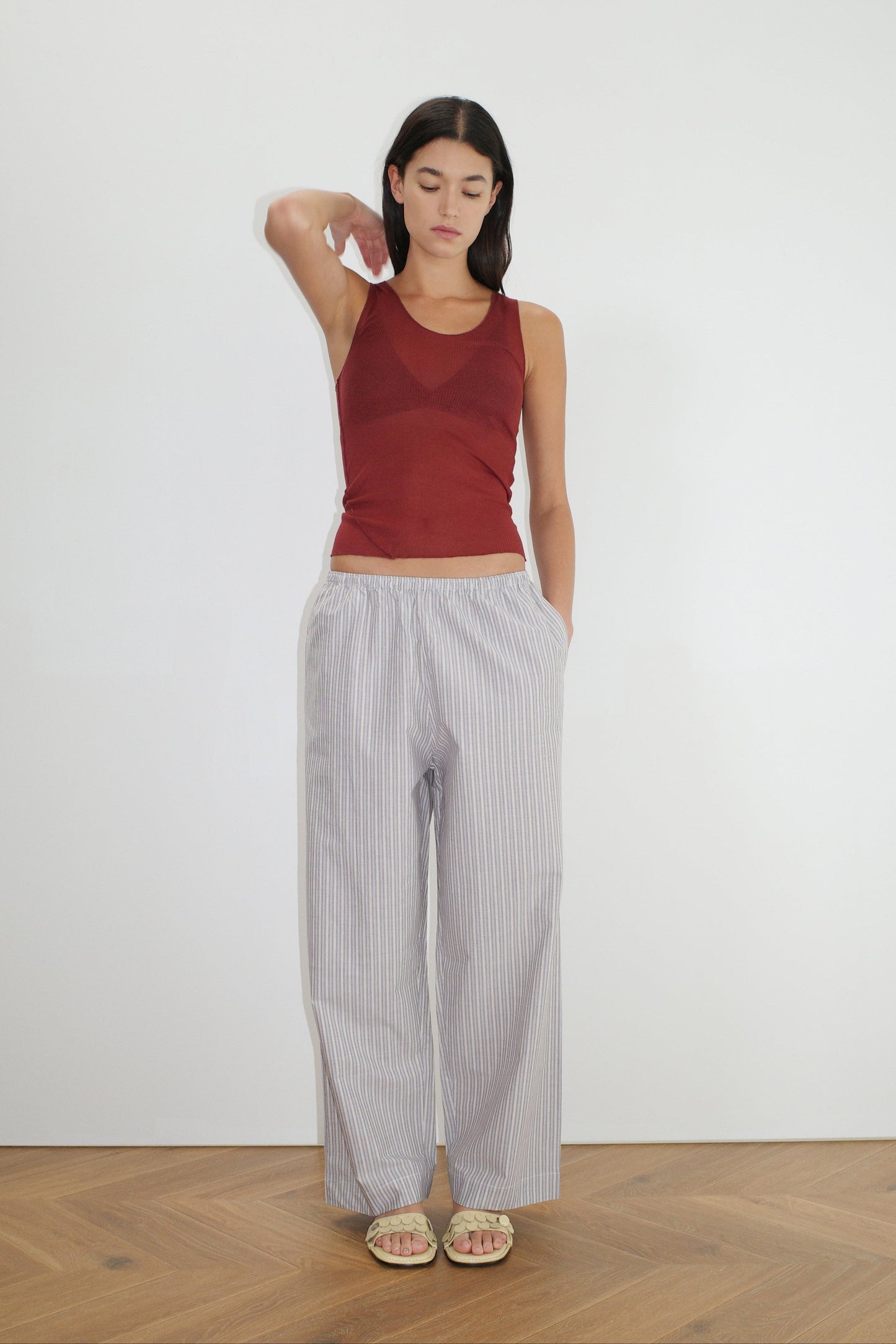 A woman stands indoors by a white wall, styling the Ease Trouser—loose light gray striped pants—with a sleeveless maroon top and sandals. One hand is in her pocket, the other raised near her head.