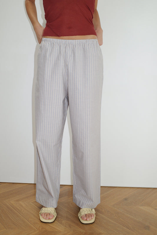 A person in Deiji Studios' Ease Trouser - Seas Stripe, a sleeveless burgundy top, and beige sandals stands on a wooden floor against a plain white wall, hands casually tucked in their pockets.