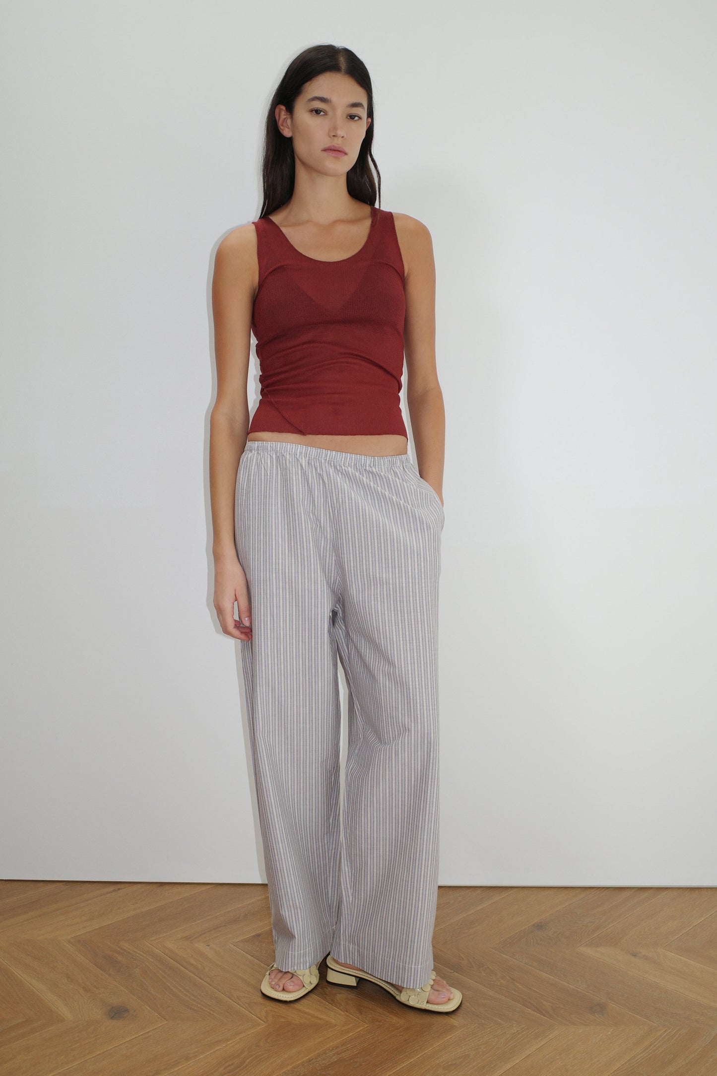 A woman with long dark hair stands against a white wall wearing a sleeveless maroon top, beige flip-flops, and Deiji Studios' Ease Trouser - Seas Stripe in light gray and white organic cotton stripes, one hand in her pocket.