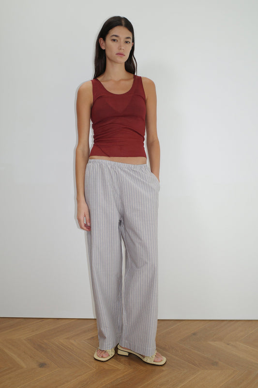 A woman with long dark hair stands against a white wall wearing a sleeveless maroon top, beige flip-flops, and Deiji Studios' Ease Trouser - Seas Stripe in light gray and white organic cotton stripes, one hand in her pocket.