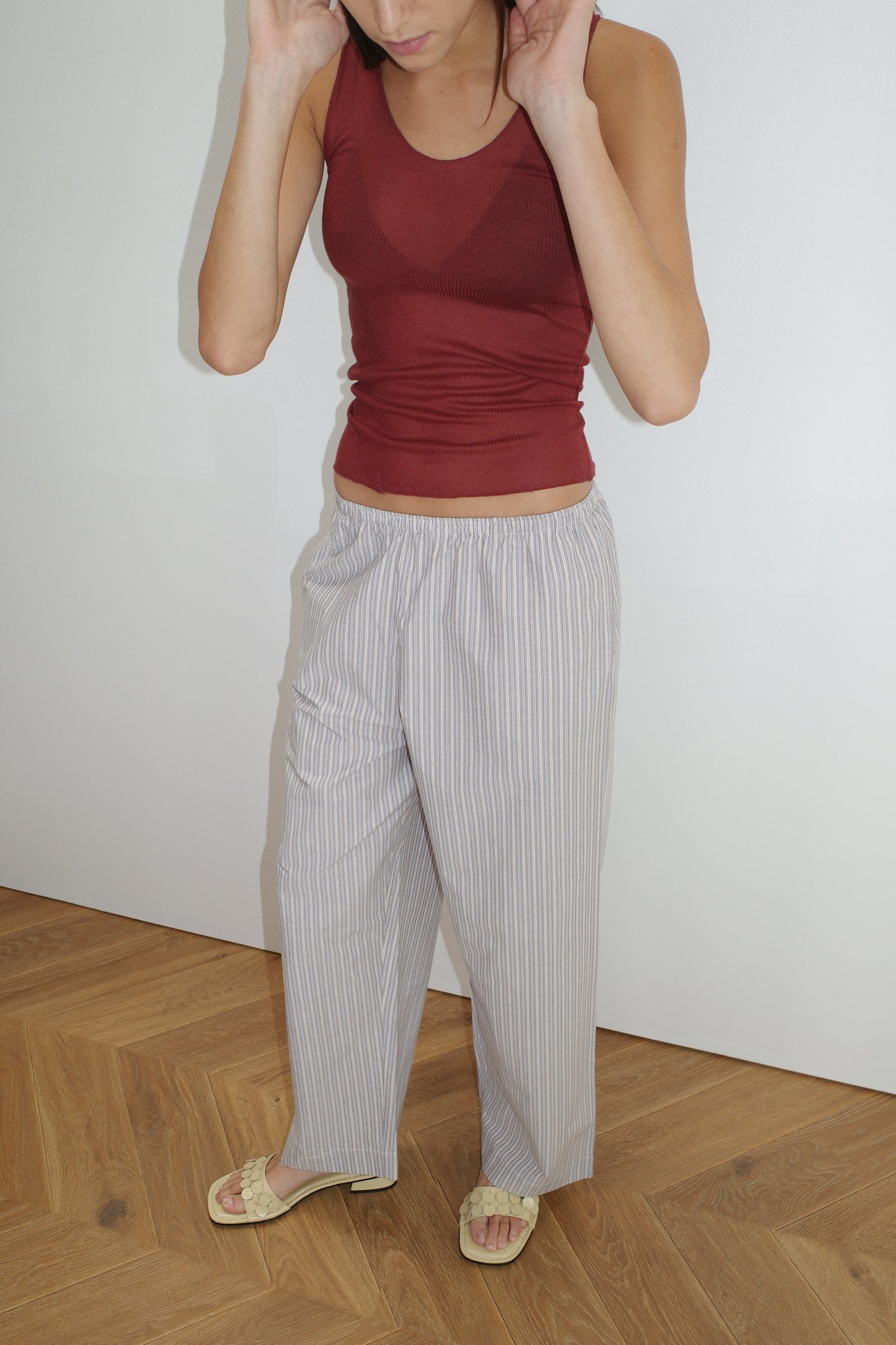 Wearing a sleeveless maroon top, Ease Trouser - Seas Stripe by Deiji Studios in organic cotton, and yellow sandals, a person stands on a wooden floor against a plain white wall, adjusting their hair with both hands.
