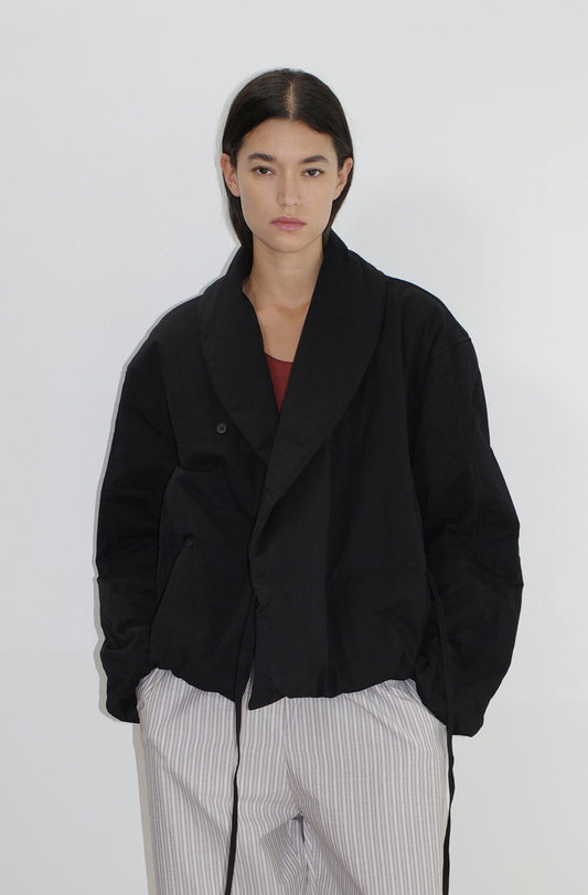 A young woman with long dark hair stands against a white background, wearing the Bubble Coat - Black by 0 over a maroon top and light gray striped pants, looking at the camera with a neutral expression.