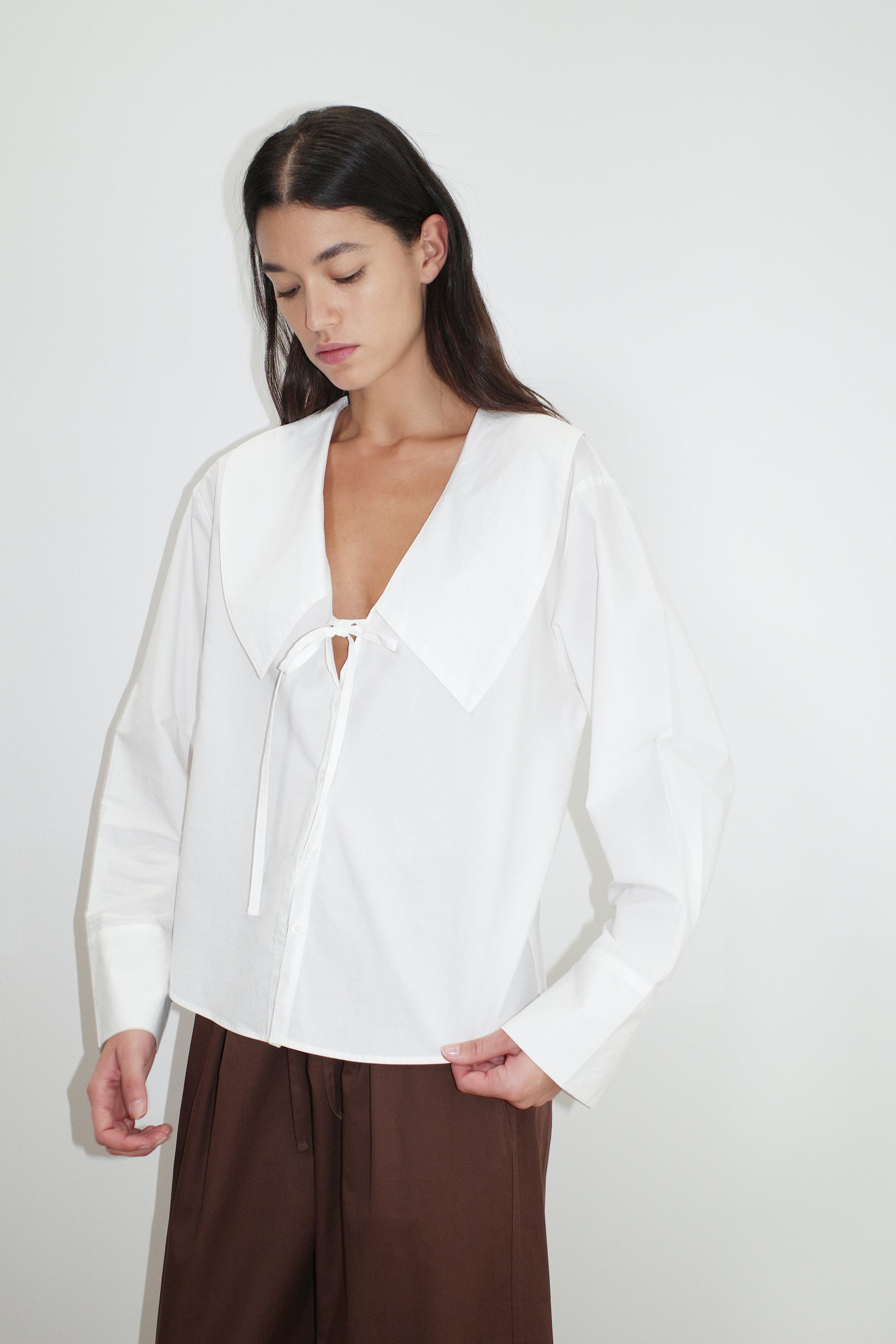 female model wears the collared tie shirt by Deiji Studios in white


a structured shirt features an oversized collar and cuff, tie at front collar opening, dropped shoulder and elongated sleeves in a boxy fit


made from 100% GOTS certified organic cotton poplin

paired with a brown cotton trouser