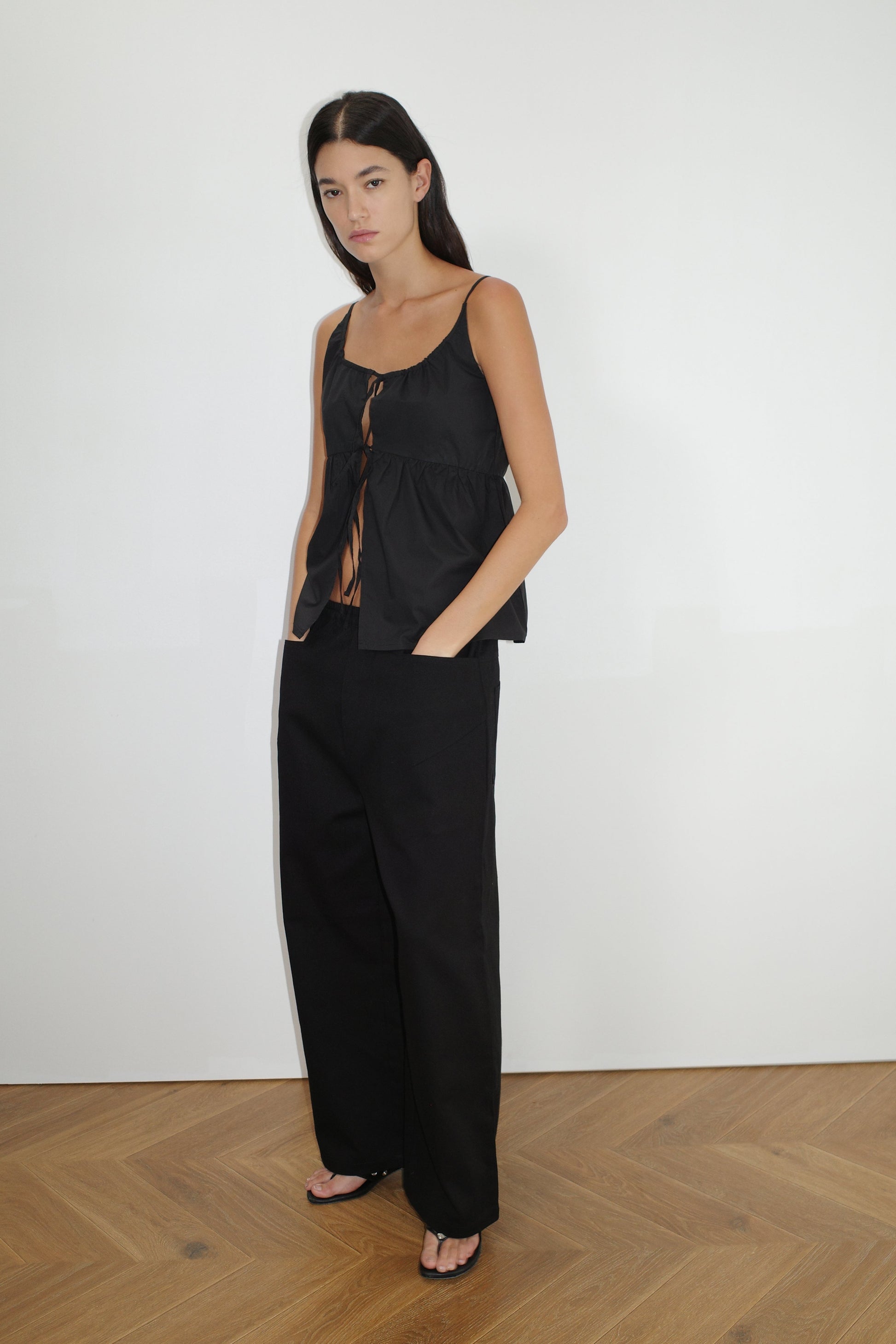 A woman with long dark hair stands on a wooden floor against a white wall, wearing Deiji Studios Square Pocket Pant in black, paired with a sleeveless black tied top and black sandals.