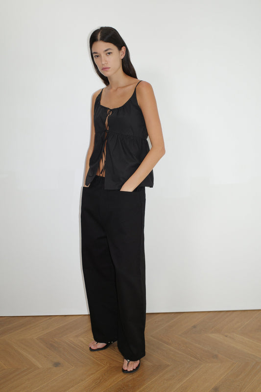A woman with long dark hair stands on a wooden floor against a white wall, wearing Deiji Studios Square Pocket Pant in black, paired with a sleeveless black tied top and black sandals.