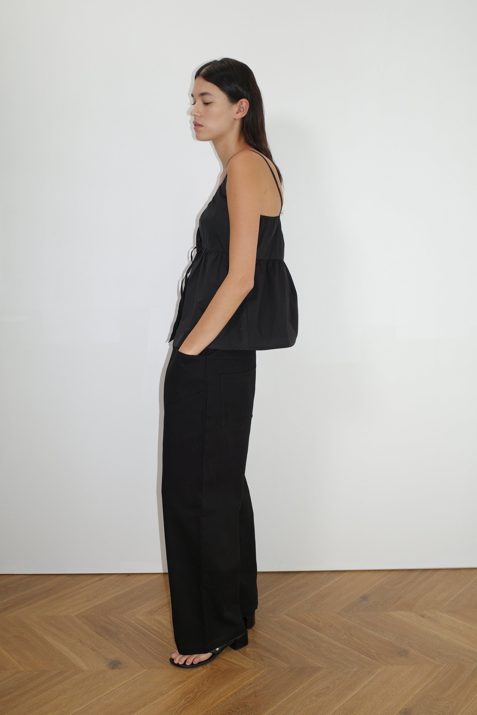 A woman with long dark hair stands in profile against a white wall, wearing 0’s Square Pocket Pant - Black, an organic cotton twill wide-leg pant with square pockets, and a sleeveless black peplum top. Her hands are in her pockets.