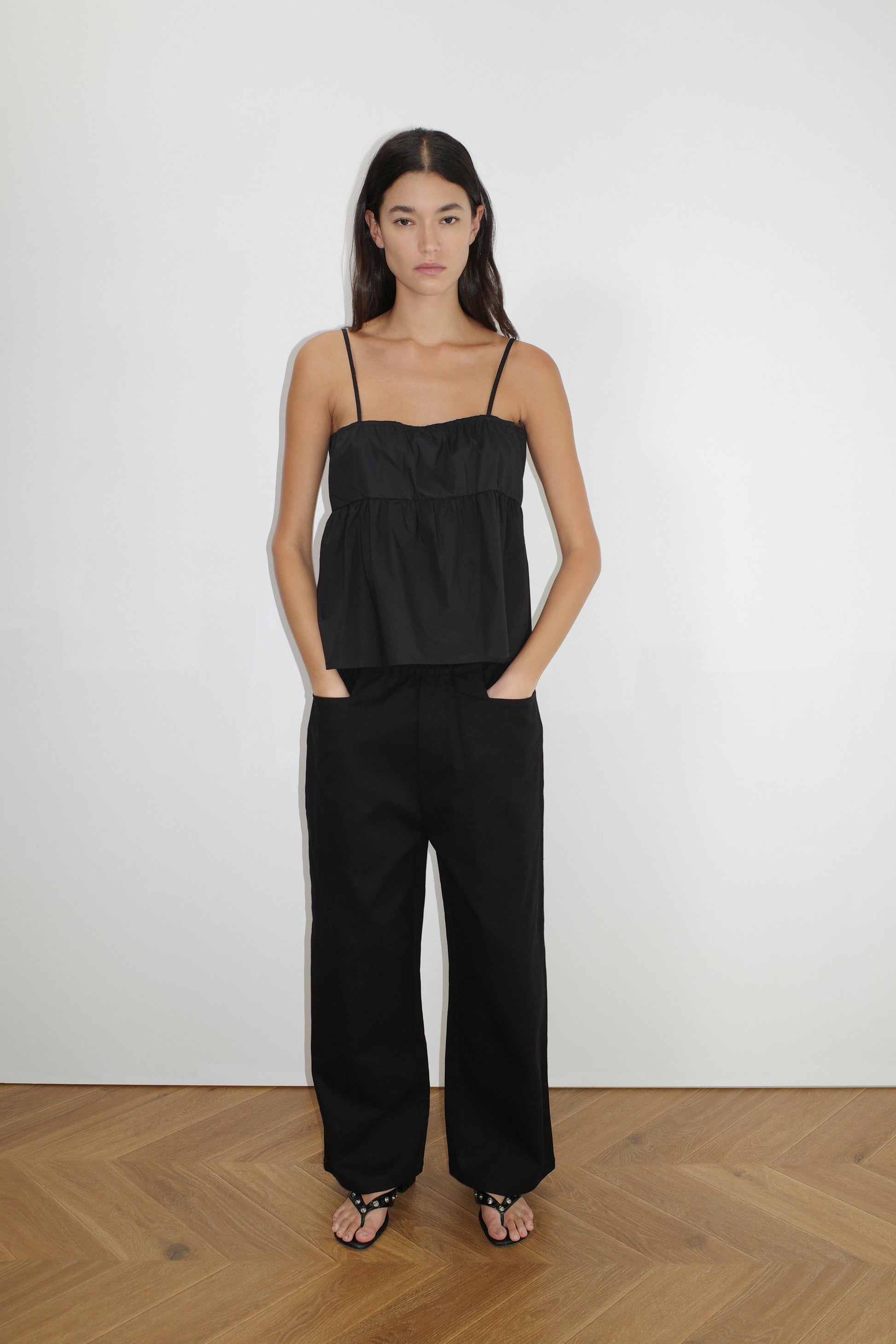 A woman with long dark hair stands against a white wall wearing the 0 Square Pocket Pant - Black in a wide-leg silhouette, paired with a loose black camisole top and black sandals, hands in her pockets on the wooden floor.