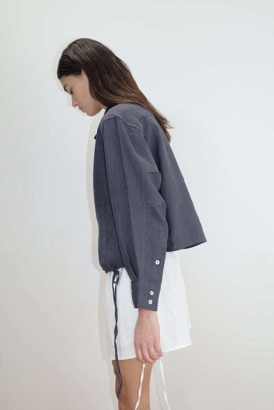 A woman with long brown hair stands sideways against a white background, wearing a loose-fitting, dark gray Hem Shirt with buttoned cuffs and white shorts. Her head is tilted slightly downward.