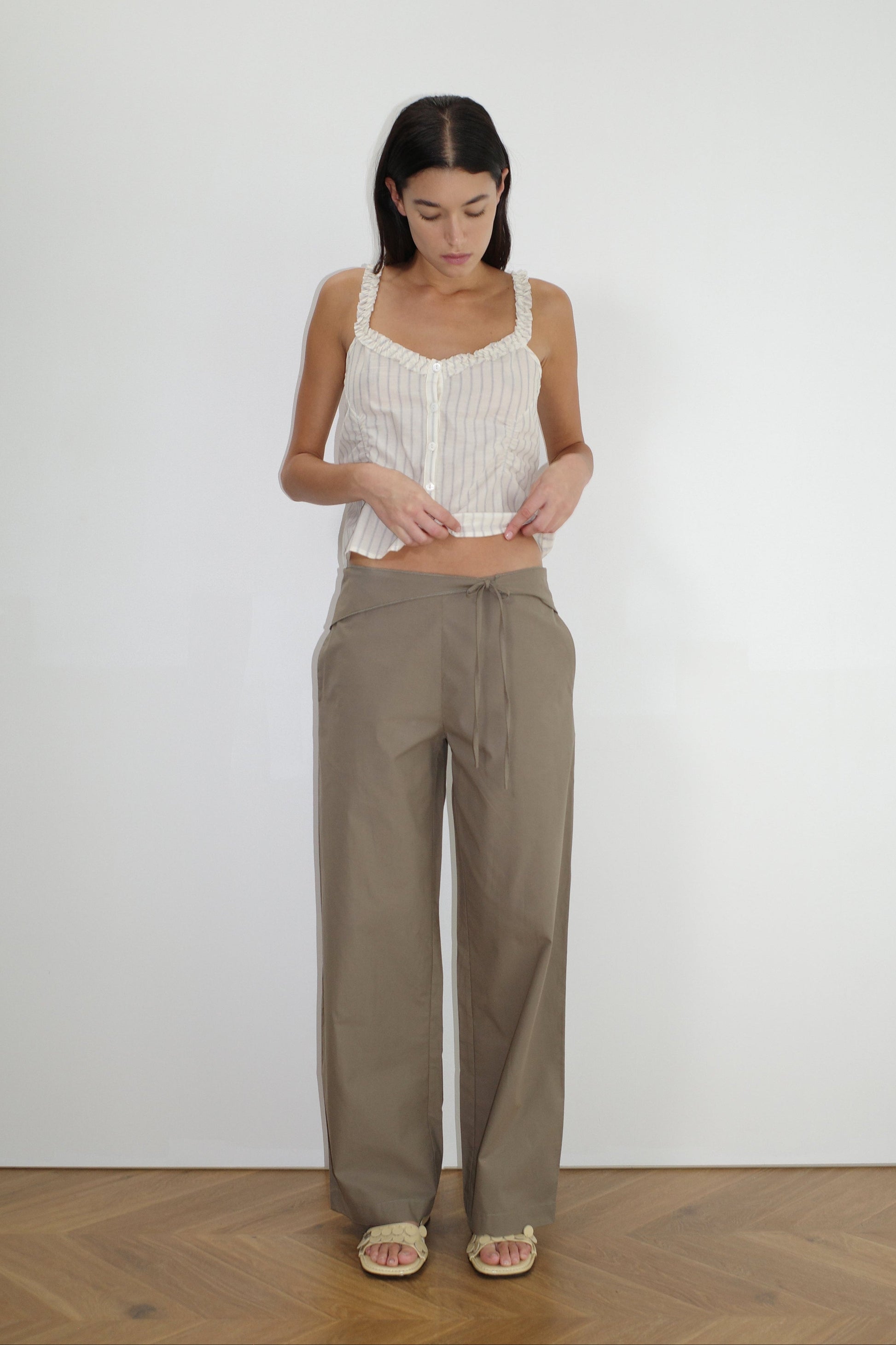 A woman stands indoors on a wooden floor, wearing the Deiji Studios Ruffle Button Top - Raquette Stripe with beige wide-leg pants and open-toed sandals, looking down against a plain white wall.