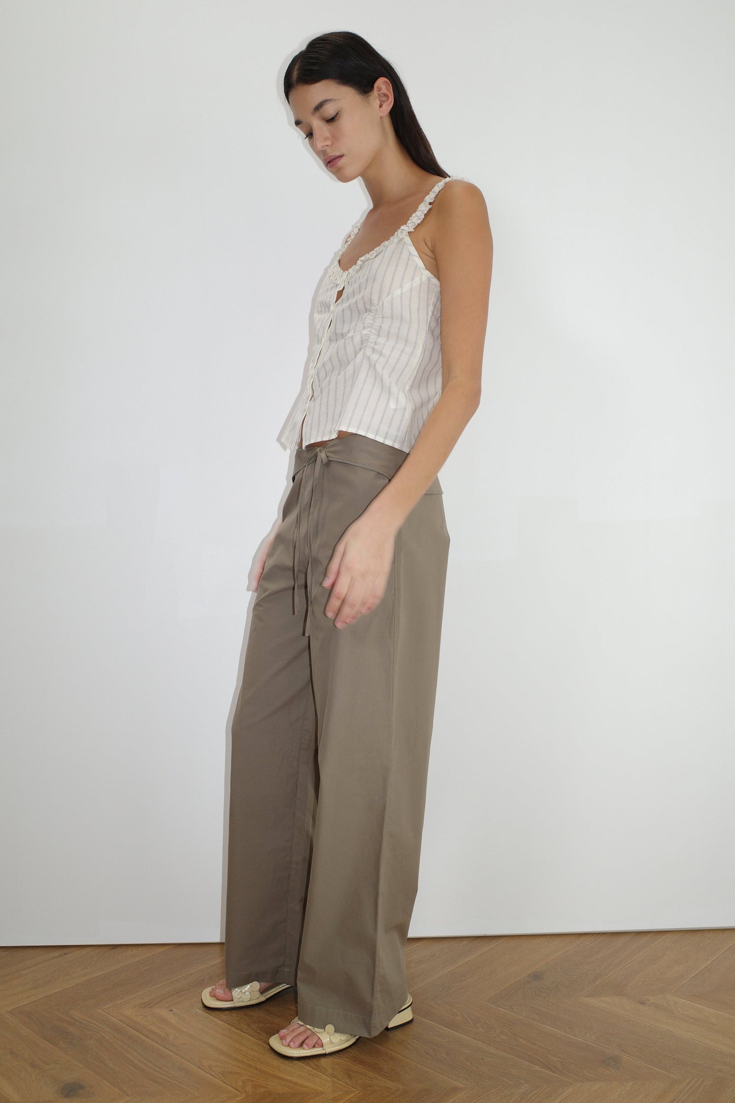 A woman stands indoors against a plain white wall, wearing the Ruffle Button Top - Raquette Stripe with wide-legged khaki pants. She has long dark hair, flat sandals, and stands on a wooden floor.