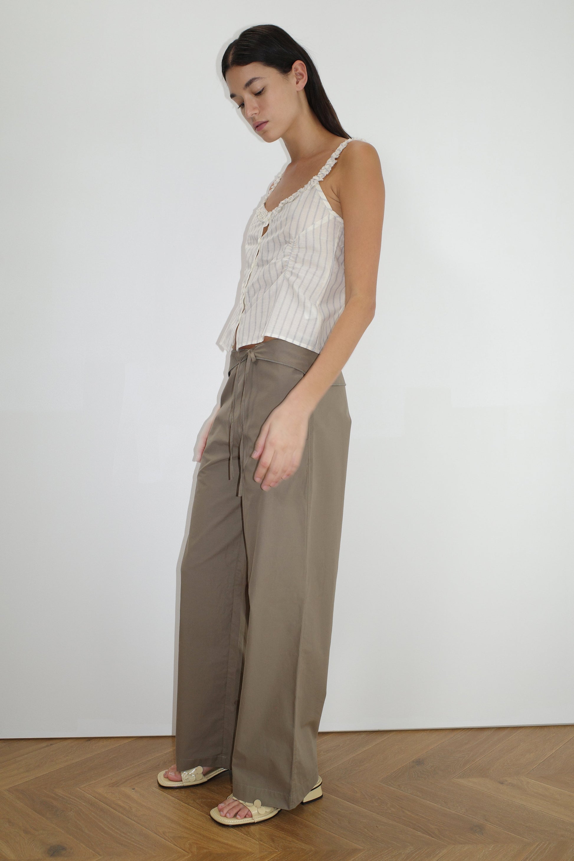 A woman stands indoors against a plain white wall, wearing the Ruffle Button Top - Raquette Stripe with wide-legged khaki pants. She has long dark hair, flat sandals, and stands on a wooden floor.
