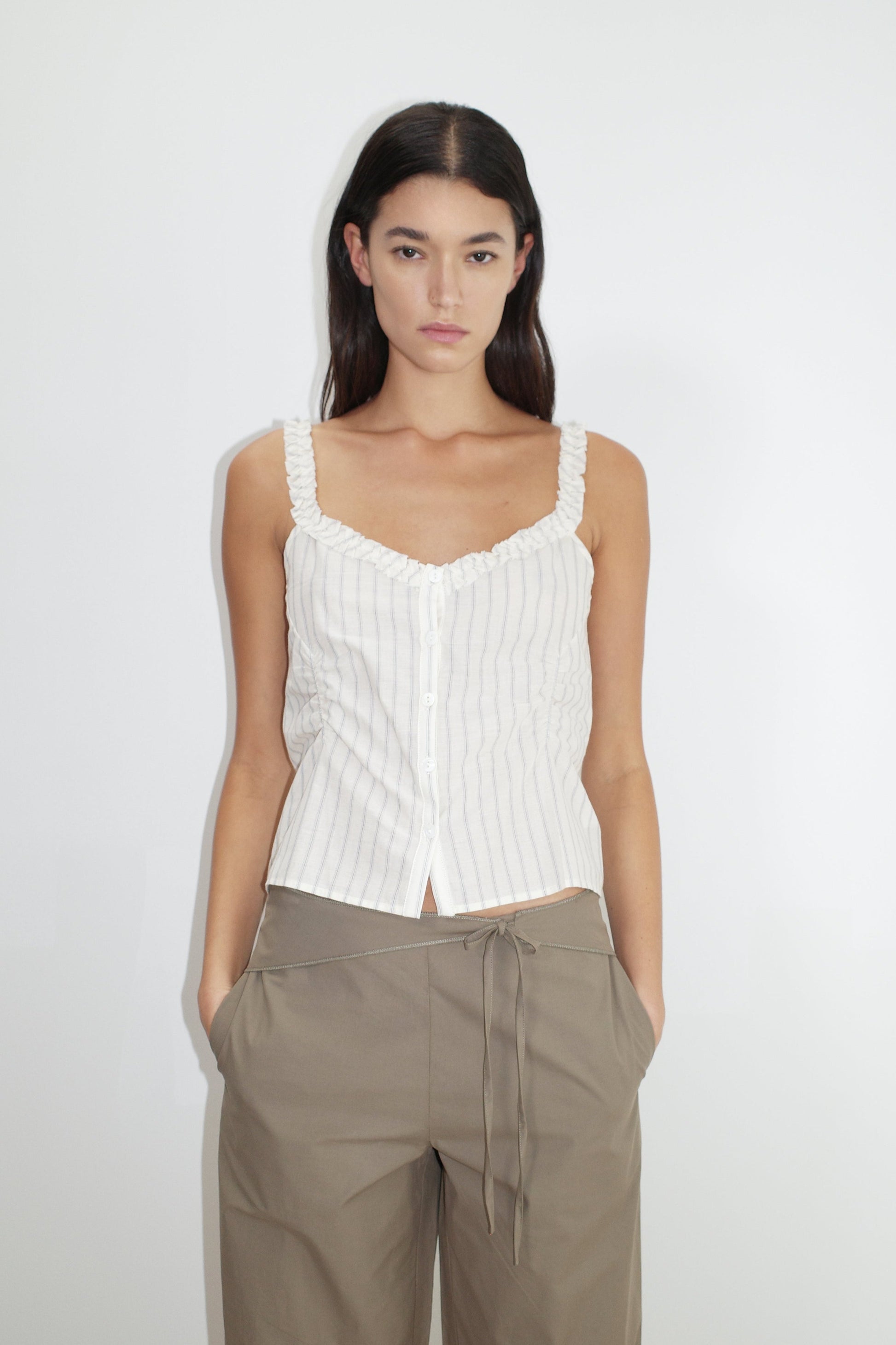 A woman with long dark hair wears a white, sleeveless, button-up striped top and Deiji Studios' Belted Pant - Clay, standing against a plain white background with her hands in her pockets.