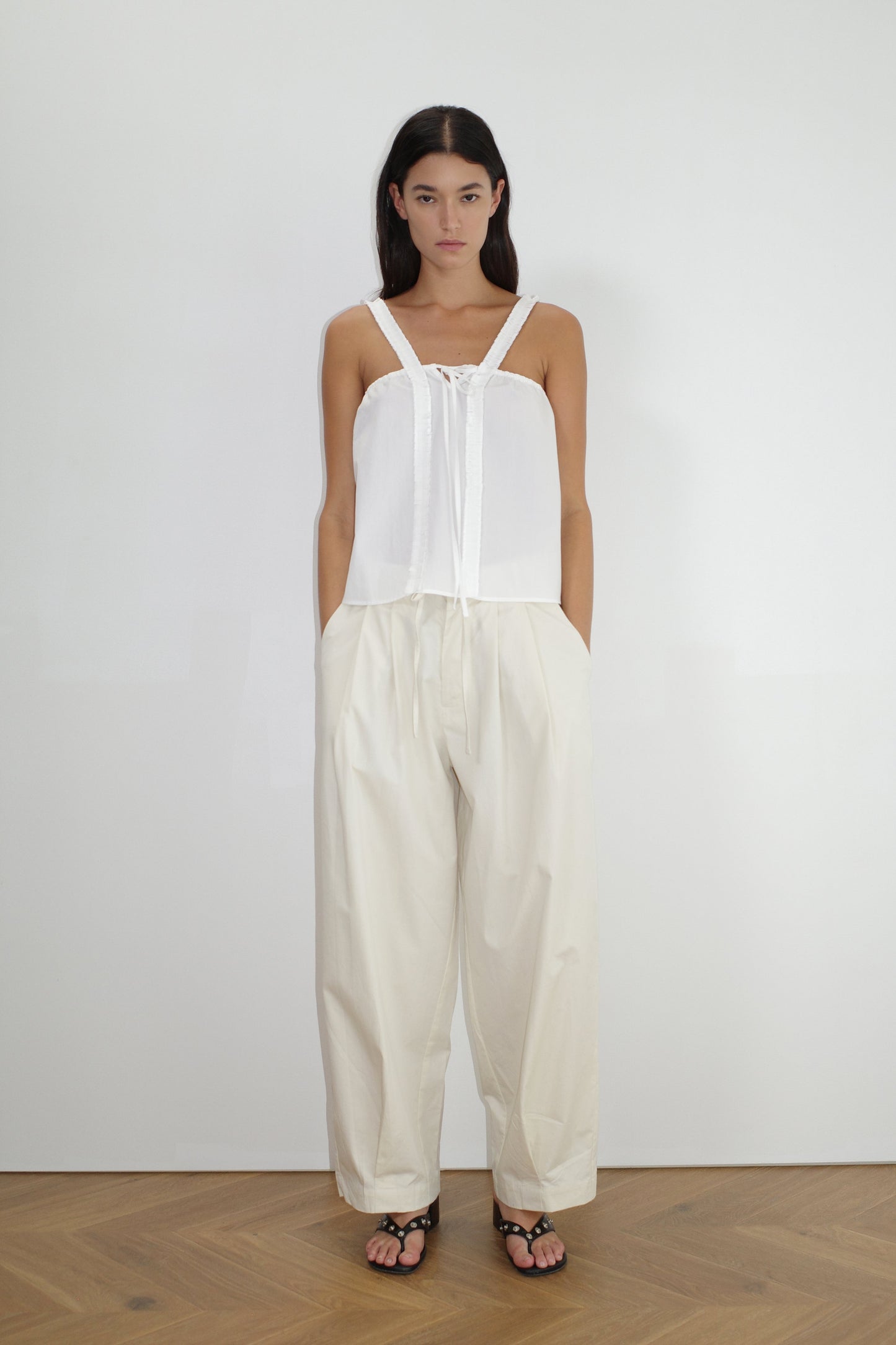 female model wears the turn pant in off white, a relaxed tailored pant features soft pleating at the front, buttoned waistband and adjustable inner drawcord. designed with hidden side seam pockets and pockets at the back, made from 100% GOTS certified organic cotton paired with a white top