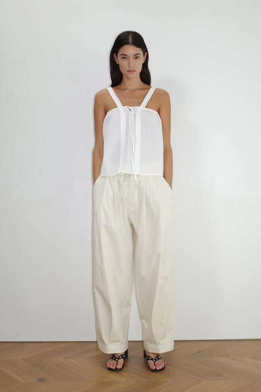 female model wears the turn pant in off white, a relaxed tailored pant features soft pleating at the front, buttoned waistband and adjustable inner drawcord. designed with hidden side seam pockets and pockets at the back, made from 100% GOTS certified organic cotton paired with a white top