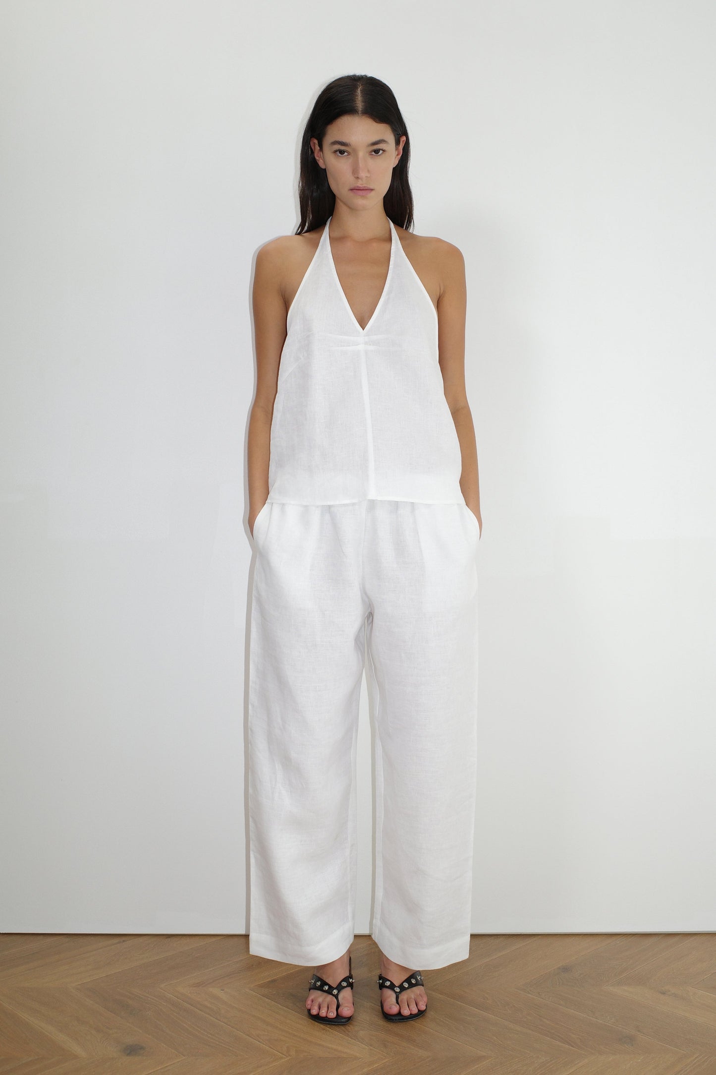 A woman with long dark hair stands indoors on a wooden floor wearing a sleeveless white halter top and the Linen Ease Trouser - White by Deiji Studios, paired with black sandals and her hands in the pockets.