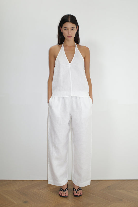 A woman with long dark hair stands indoors on a wooden floor wearing a sleeveless white halter top and the Linen Ease Trouser - White by Deiji Studios, paired with black sandals and her hands in the pockets.