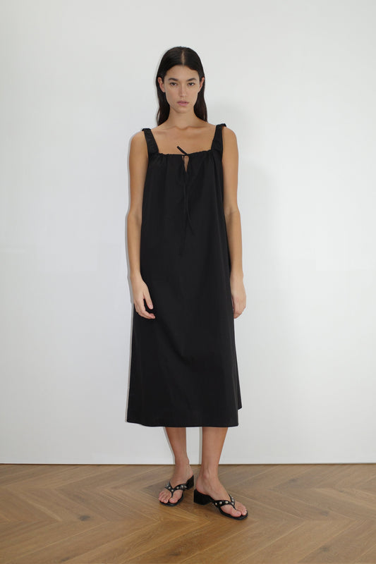 A woman with long dark hair stands indoors on a wooden floor, wearing The Paper Dress - Black by Deiji Studios—a loose, sleeveless maxi dress with a square neckline and bow tie detail—paired with black sandals, facing forward against a plain white wall.