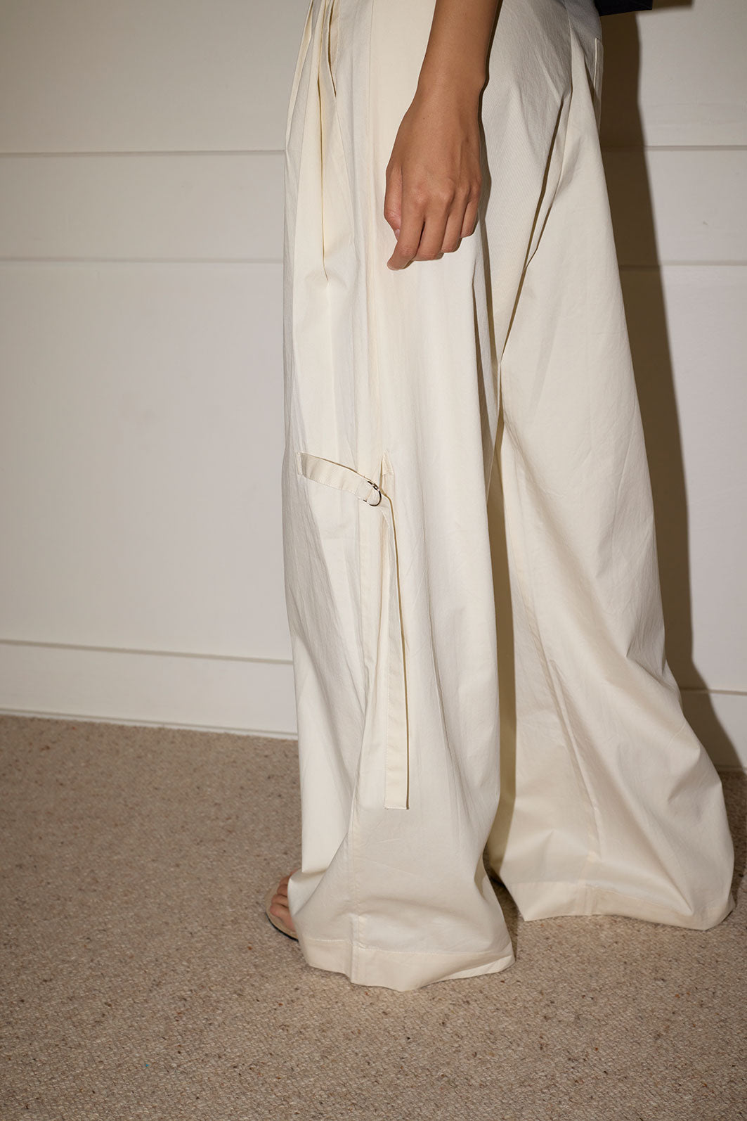 Someone stands indoors on a beige carpet, wearing Deiji Studios' Tab Pant in Off White organic cotton. Their arm rests by their side, and behind them is a white wall with decorative molding.