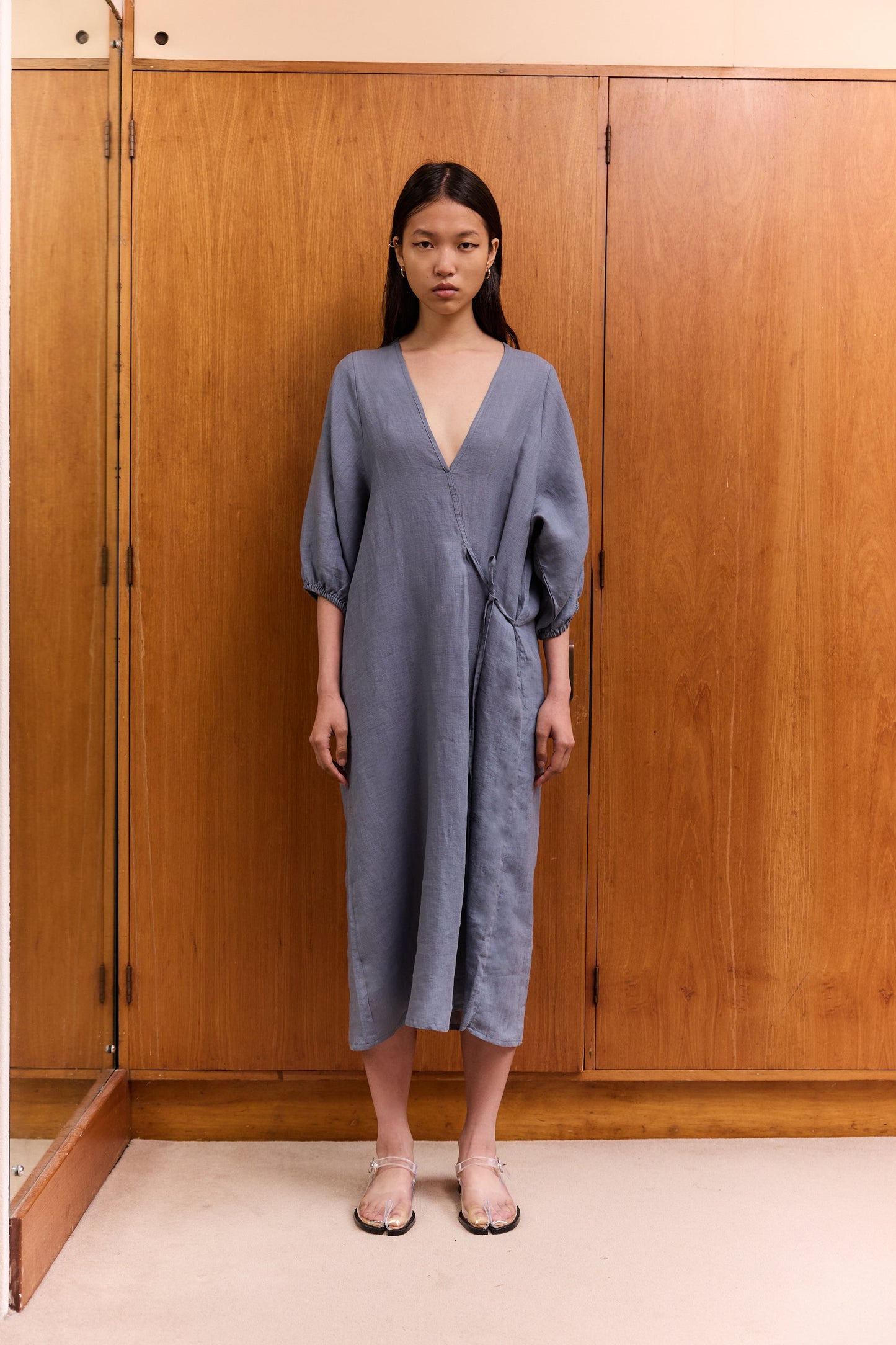 A woman stands indoors by wooden cabinets, wearing the Crease Bind Dress—a loose-fitting, long-sleeved blue wrap dress with a deep V-neck—paired with light sandals. She looks at the camera with a neutral expression.