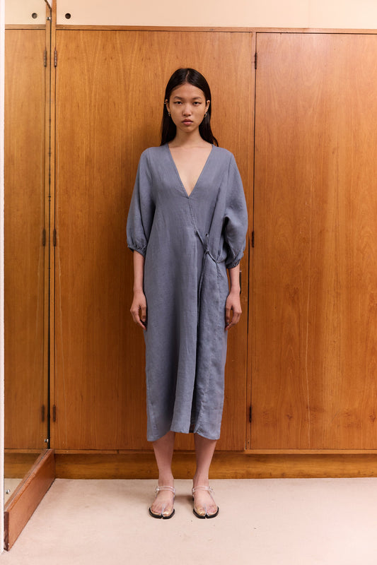 A woman stands indoors by wooden cabinets, wearing the Crease Bind Dress—a loose-fitting, long-sleeved blue wrap dress with a deep V-neck—paired with light sandals. She looks at the camera with a neutral expression.